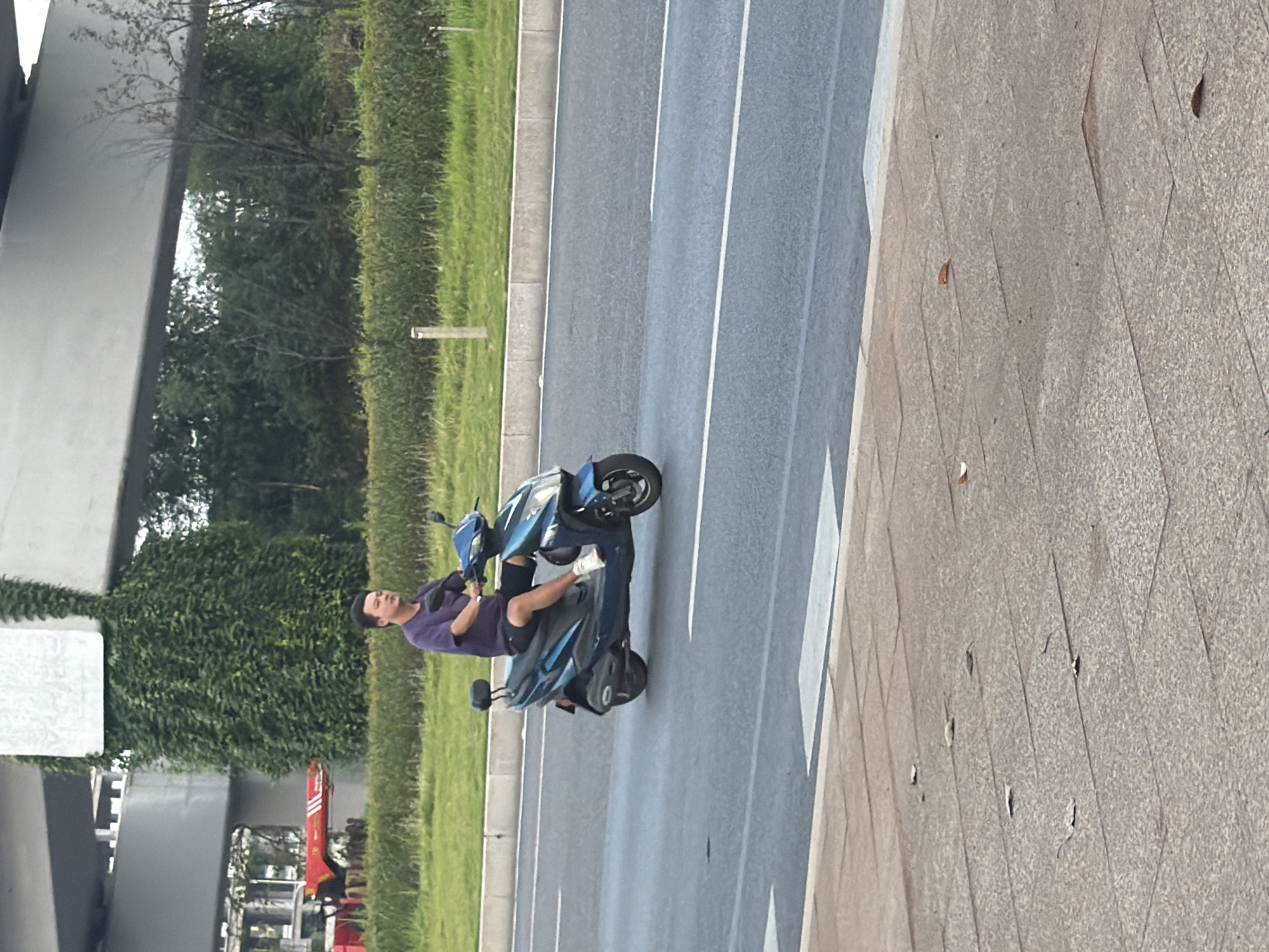 A man wearing a purple t-shirt and dark shorts is riding a blue electric scooter on a multi-lane asphalt road. The scooter has a black seat and a small black backrest. The man is looking forward and appears to be steering the scooter. The road is bordered by a grassy median with a concrete curb and a metal guardrail. In the background, there are trees and vegetation. Above the trees, a large concrete overpass is visible, partially covered in green vines. On the left side of the image, a red fire truck is parked behind the guardrail. The foreground shows a tiled sidewalk with scattered leaves. The scene appears to be in the city of Jiangjin, China.