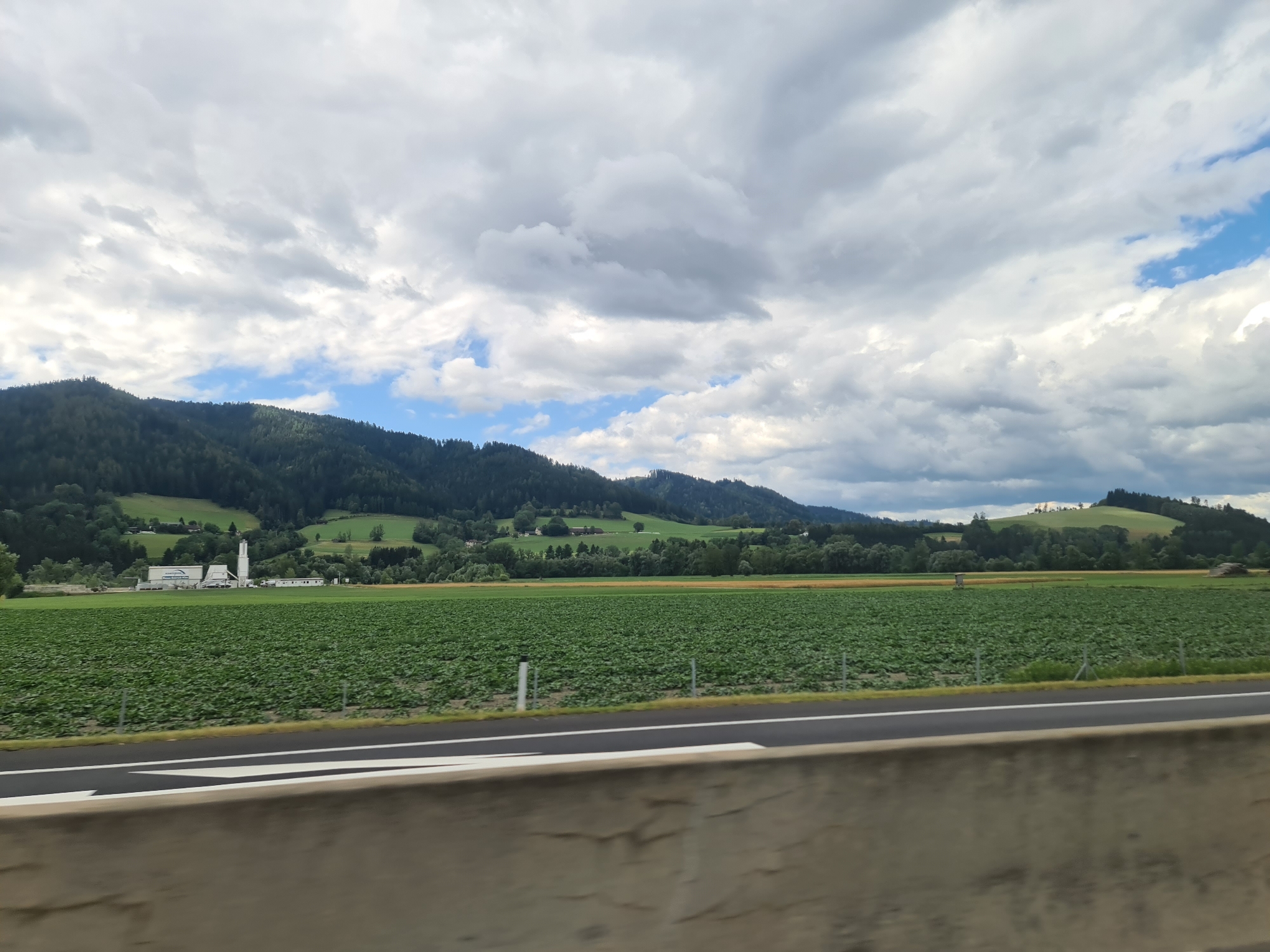 An expansive, wide-angle outdoor photograph captures a scenic rural landscape under a dramatic, cloudy sky. The foreground features a concrete barrier and a paved road with white and black lane markings, suggesting a highway or a similar thoroughfare. Beyond the road, a vast, flat field of lush green crops stretches towards a range of rolling, tree-covered hills in the mid-ground. A cluster of white buildings with tall structures, possibly industrial or agricultural facilities, is visible on the left side of the field. The background is dominated by a chain of densely forested mountains, their peaks partially obscured by the heavy, grey clouds that fill the upper portion of the frame. Patches of blue sky peek through the clouds, hinting at the possibility of clearing weather. The overall impression is one of a tranquil, undeveloped rural area. The location is identified as Kaisersberg, Austria.