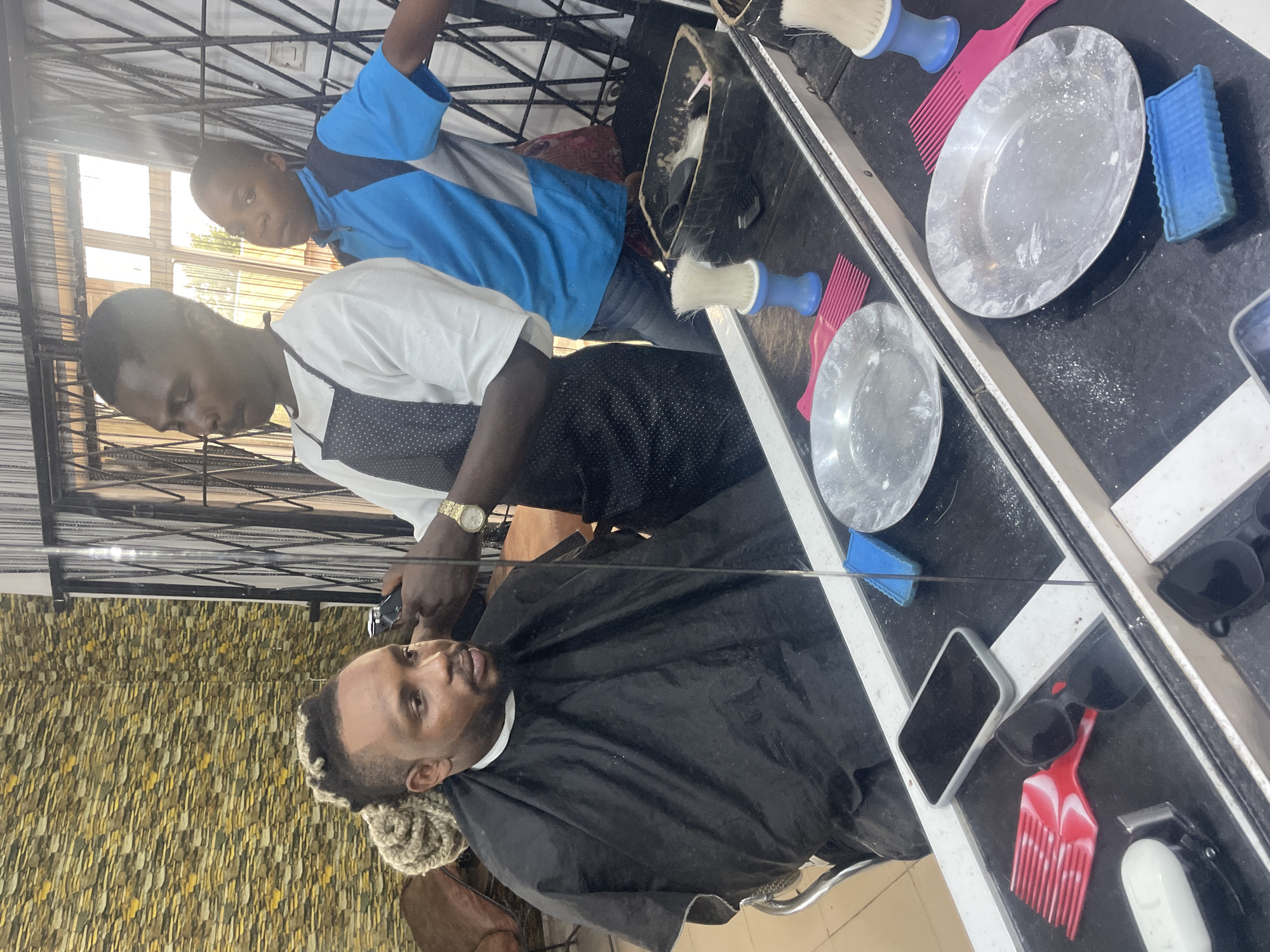 A man is receiving a haircut in a barber shop. The barber, dressed in a white shirt with a black vest, is using an electric clipper on the client. The client is draped in a black cape and has blonde highlights. Another man in a blue shirt is standing behind the barber. The barber shop has a black and white counter with barber supplies such as brushes, combs, and bowls on top. The walls are decorated with a textured brown and beige design. The location is in Jos, Nigeria.