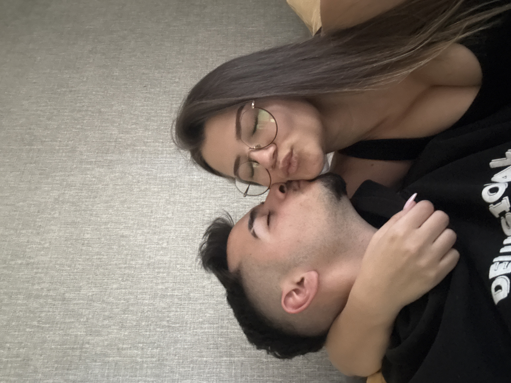 A woman with brown hair and glasses kisses a man on the cheek. The man has dark hair and stubble. The background is a gray surface. The city of Sofia, Bulgaria is not identifiable in the image.