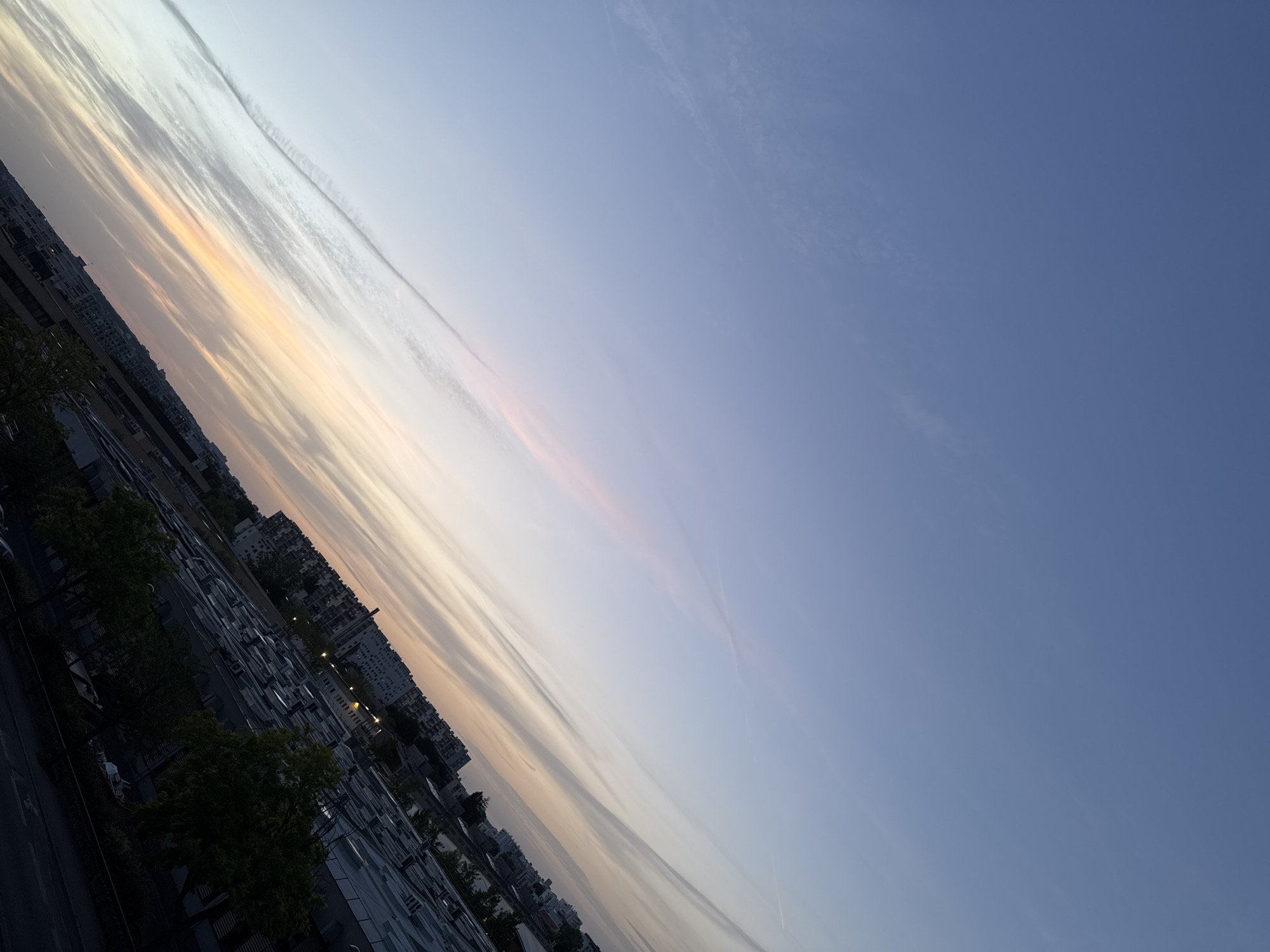 The image shows a cityscape at dusk, with the sky displaying a gradient of colors from deep blue at the top to soft orange and pink near the horizon. Long, thin clouds stretch across the sky, some appearing as contrails. Below the sky, a dense urban landscape is visible, characterized by a mix of residential buildings and commercial or industrial structures with flat roofs and numerous air conditioning units. Trees with dark green foliage are scattered throughout the foreground and midground, casting shadows. The buildings appear to be of various heights and architectural styles, with some featuring balconies and windows. Streetlights are beginning to illuminate in the distance. The scene is captured from a high vantage point, possibly a rooftop, looking out over the city. The overall impression is one of a tranquil evening in an urban environment.