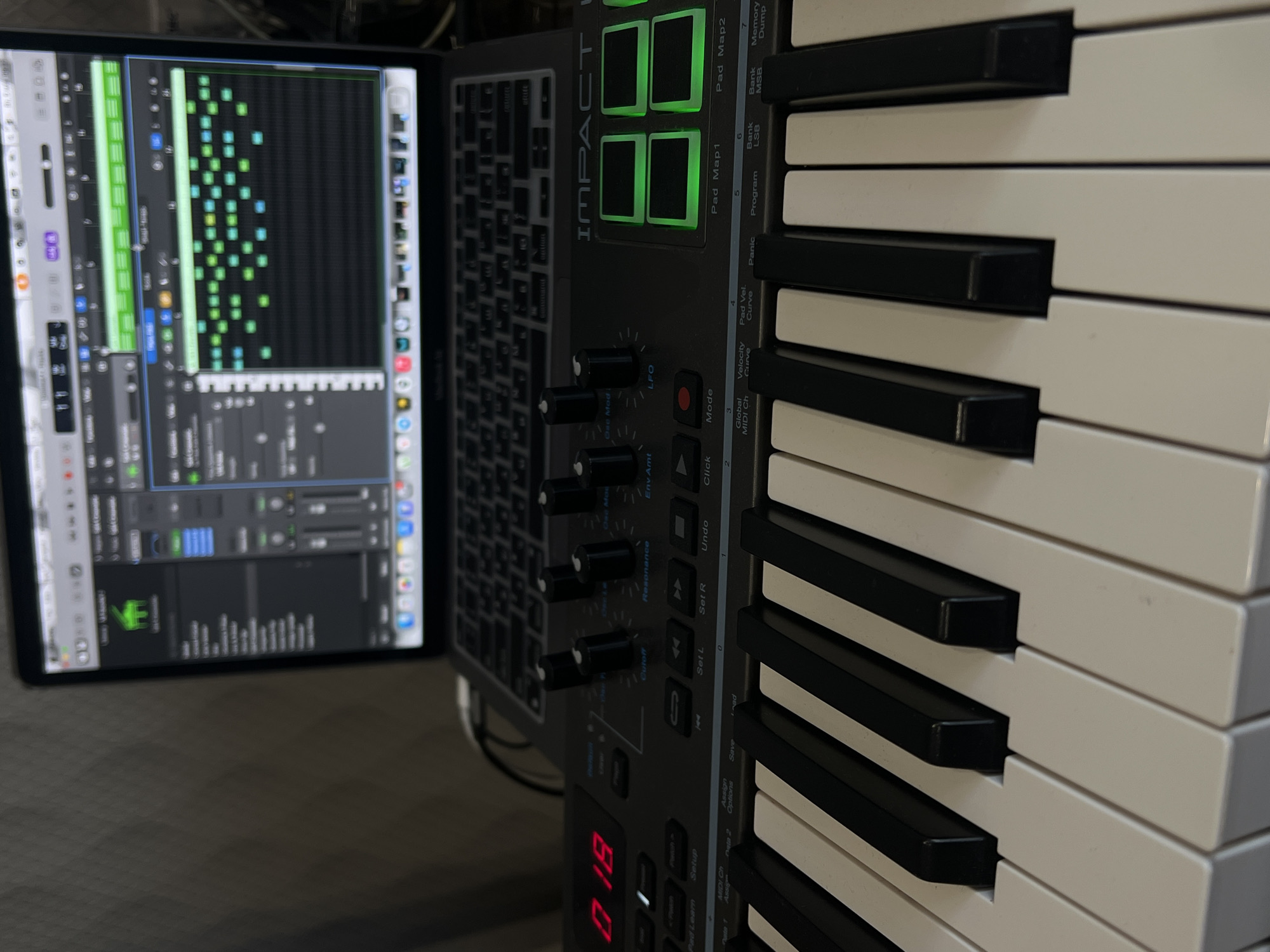The image depicts a close-up view of a digital music production setup. The scene focuses on a MIDI keyboard controller with black and white keys, knobs, and illuminated buttons. A laptop screen displays digital audio workstation (DAW) software with a piano roll view. The equipment is arranged on a neutral-colored surface. There is no information available in the image to determine if the photo was taken in Krasnodar, Russia.