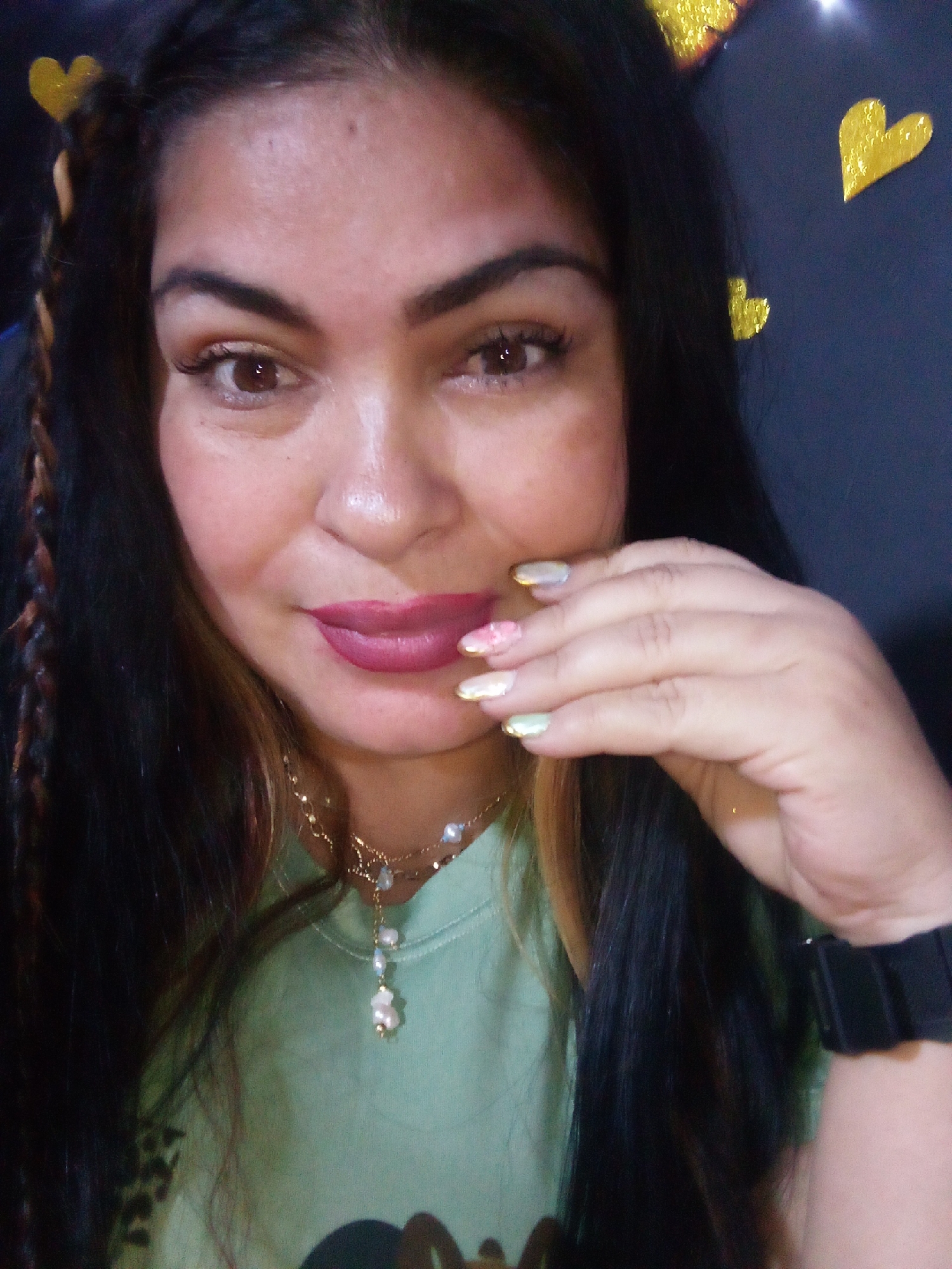 A close-up, frontal portrait depicts an adult female positioned indoors in Juan Griego, Venezuela. She has long, dark brown hair with visible lighter streaks and a thin braid near her left temple. Her eyes are dark brown, and her lips are coated with dark pink lipstick. Her left hand, displaying multi-colored manicured nails with pastel hues and gold accents, is held near her chin. She wears a light green long-sleeved top, a gold-toned necklace with dangling bead charms, and a dark digital watch on her left wrist. The dark background features multiple yellow, glittery heart-shaped decorations affixed to it, with light reflections present in the upper right.