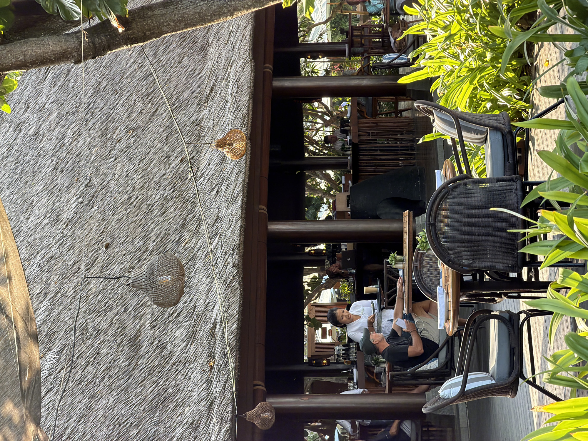 The image depicts an outdoor dining area of a restaurant with a thatched roof. In the foreground, there are several rattan chairs and a round wooden table with a white plate. In the midground, a waiter in a white shirt is serving a man in a black t-shirt and a baseball cap who is seated at a table. Behind them, a bar area is visible with bottles and glasses. The background shows trees and umbrellas, suggesting a tropical location. The scene is set in Denpasar, Indonesia, likely a resort or beachside establishment given the tropical flora and open-air setup.