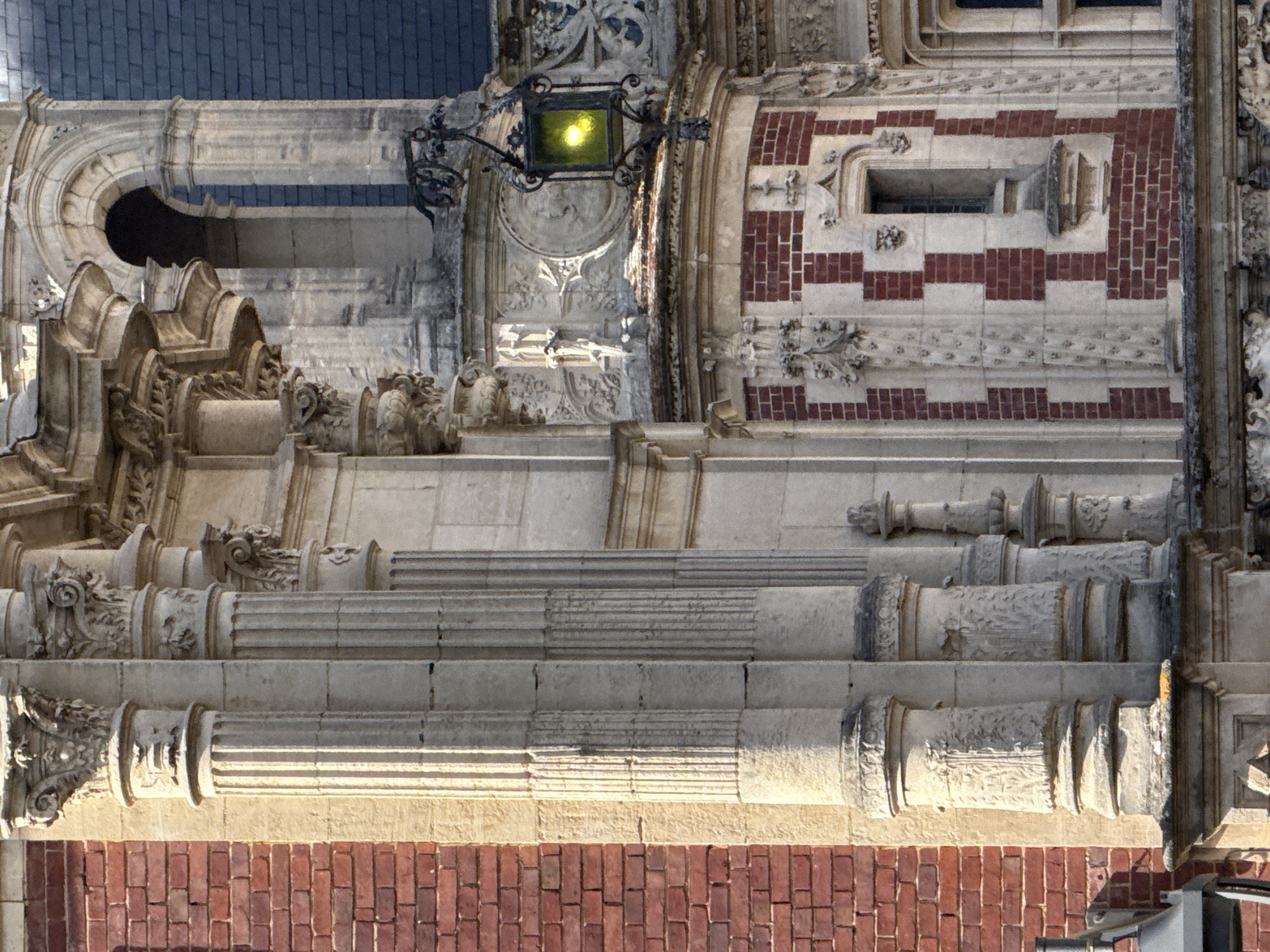 This detailed image captures a section of a highly ornate building facade, presented rotated 90 degrees clockwise. The architecture combines various materials and elaborate decorative elements, indicative of a historical or grand structure, likely located in Fécamp, France.
The top portion of the image displays a dark blue/grey tiled roof. Below this, a grand, arched stone opening, possibly part of a window or a recessed balcony, is visible, featuring intricate carvings and decorative moldings.
Towards the center of the image, an elaborate black metal lamppost is prominently featured, attached to a richly sculpted stone section. The lamppost emits a bright yellow light, even though the scene appears to be set during the daytime, with strong, angled sunlight casting deep shadows across the facade and highlighting its textures. Adjacent to this, the building's surface transitions into a striking pattern of alternating red brick and light-colored stone bands, creating a checkerboard-like design. A small, rectangular window with a stone frame is set within this patterned brickwork.
Further down the facade, multiple levels of decorative stonework are apparent. Several columns and pilasters are visible, showcasing diverse designs; some are fluted, while others are extensively carved with foliage, scrollwork, and what appear to be grotesque figures, characteristic of Gothic or Gothic Revival architectural styles. The stone exhibits signs of age and weathering in various places. The lowest visible section of the building consists of more uniform red brickwork.
No people or explicit activities are depicted in the image. The focus is entirely on the intricate craftsmanship and textural qualities of the building's exterior under clear, sunny conditions. No visible text is present.