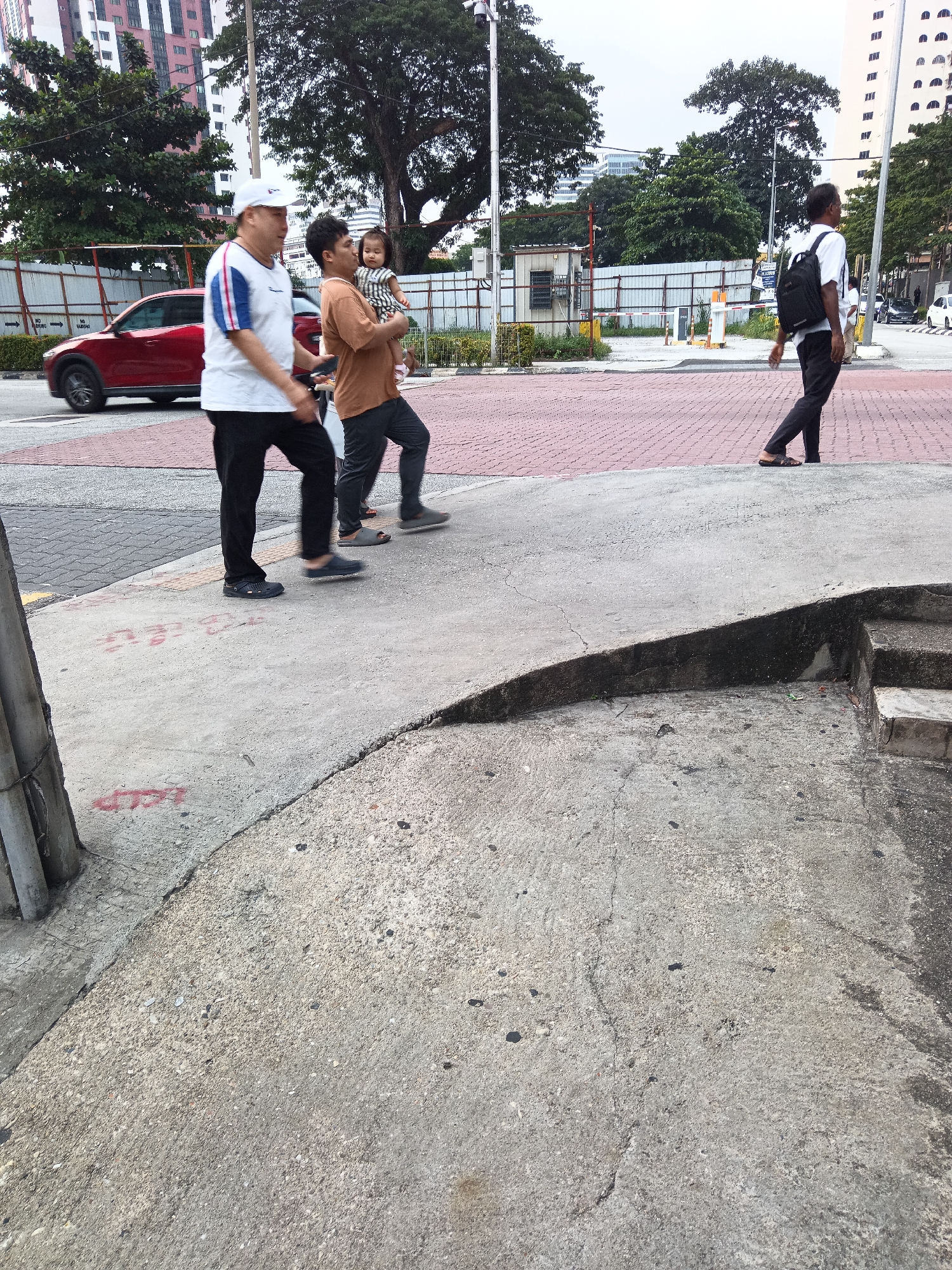 An outdoor, eye-level, medium shot captures a street scene in Kuala Lumpur, Malaysia. In the foreground, a rough concrete sidewalk dominates the lower portion of the frame, with a curved ramp leading to a raised area. To the left, a gray cylindrical post stands, showing signs of wear and possibly graffiti. Behind the sidewalk, a red-bricked pedestrian path is visible. Further back, a red car is parked on the road. Three men and a young child are walking along the pedestrian path. The man on the far left wears a white cap, a white t-shirt with blue and red accents on the sleeves, and black pants. Next to him, another man, wearing a brown t-shirt and black pants, carries a baby girl in his arms. The baby wears a striped dress. The third man, on the far right, walks away from the camera, wearing a white t-shirt, dark pants, and a black backpack. He is looking to his right. In the background, trees and tall buildings, including a pink high-rise building, indicate an urban setting. Construction barriers and fences suggest ongoing development in the area. The sky is overcast, indicating a cloudy day.