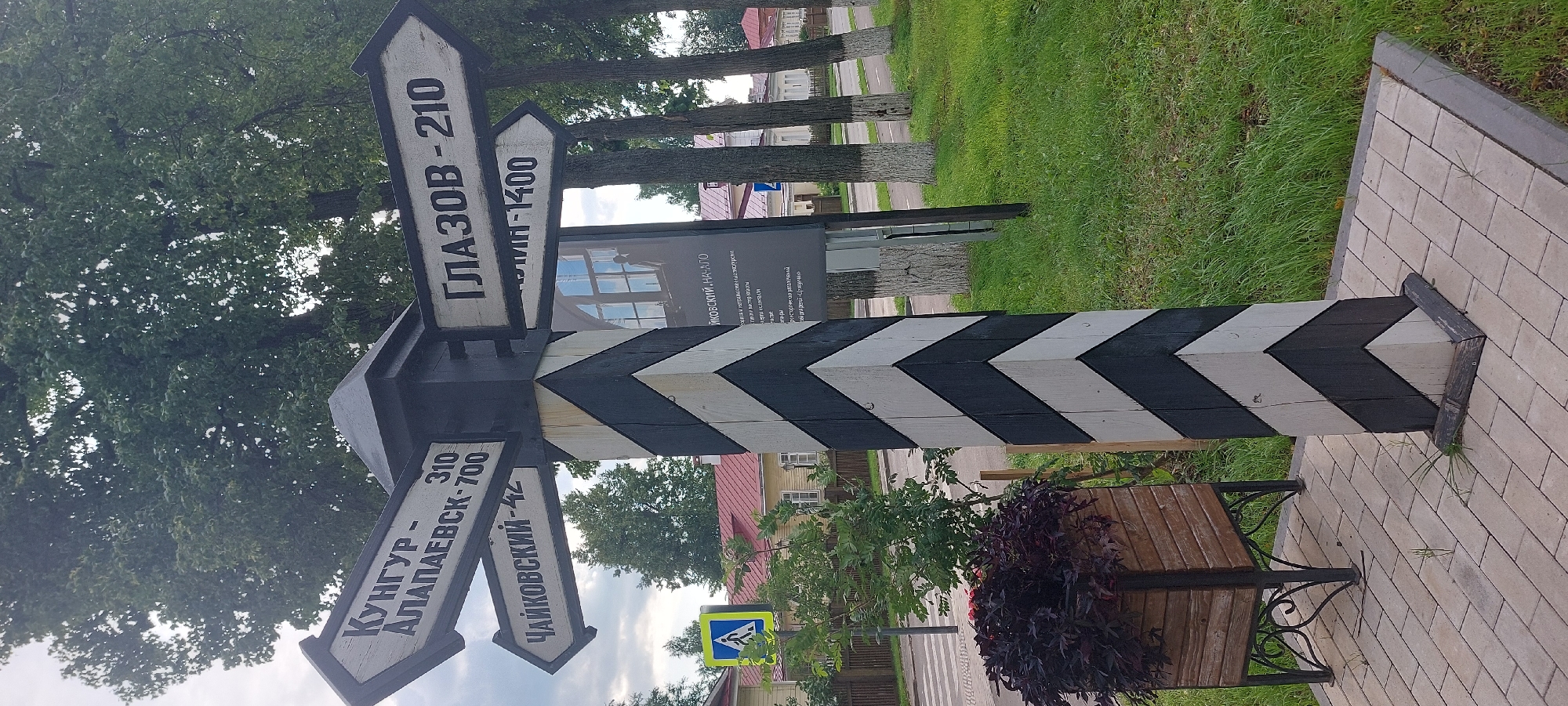 This is a medium-shot, eye-level photograph of a directional signpost in Votkinsk, Russia. The signpost is a wooden pillar painted with alternating black and white chevron patterns. It has multiple directional arrows pointing in different directions. The arrows are also painted white with black lettering. The text on the arrows indicates distances to various locations.
The arrow pointing to the left reads "КУНГТР - АЛАПАЕВСК - 700", with a smaller number "310" above it. Below this arrow, another arrow points to the left and reads "ЧАЙКОВСКИЙ - 42".
The arrow pointing to the right reads "ГЛАЗОВ - 210". Below this arrow, another arrow points to the right and reads "- 1400" which is partially obscured.
In the background, there is a tall tree with green foliage, and further back, houses with red roofs. To the left of the signpost, there is a blue and white pedestrian crossing sign. In front of the signpost, there is a wooden planter box filled with dark red foliage. The ground is paved with light-colored rectangular bricks. The scene is set outdoors on a cloudy day. A poster is attached to the signpost, displaying text in Russian.