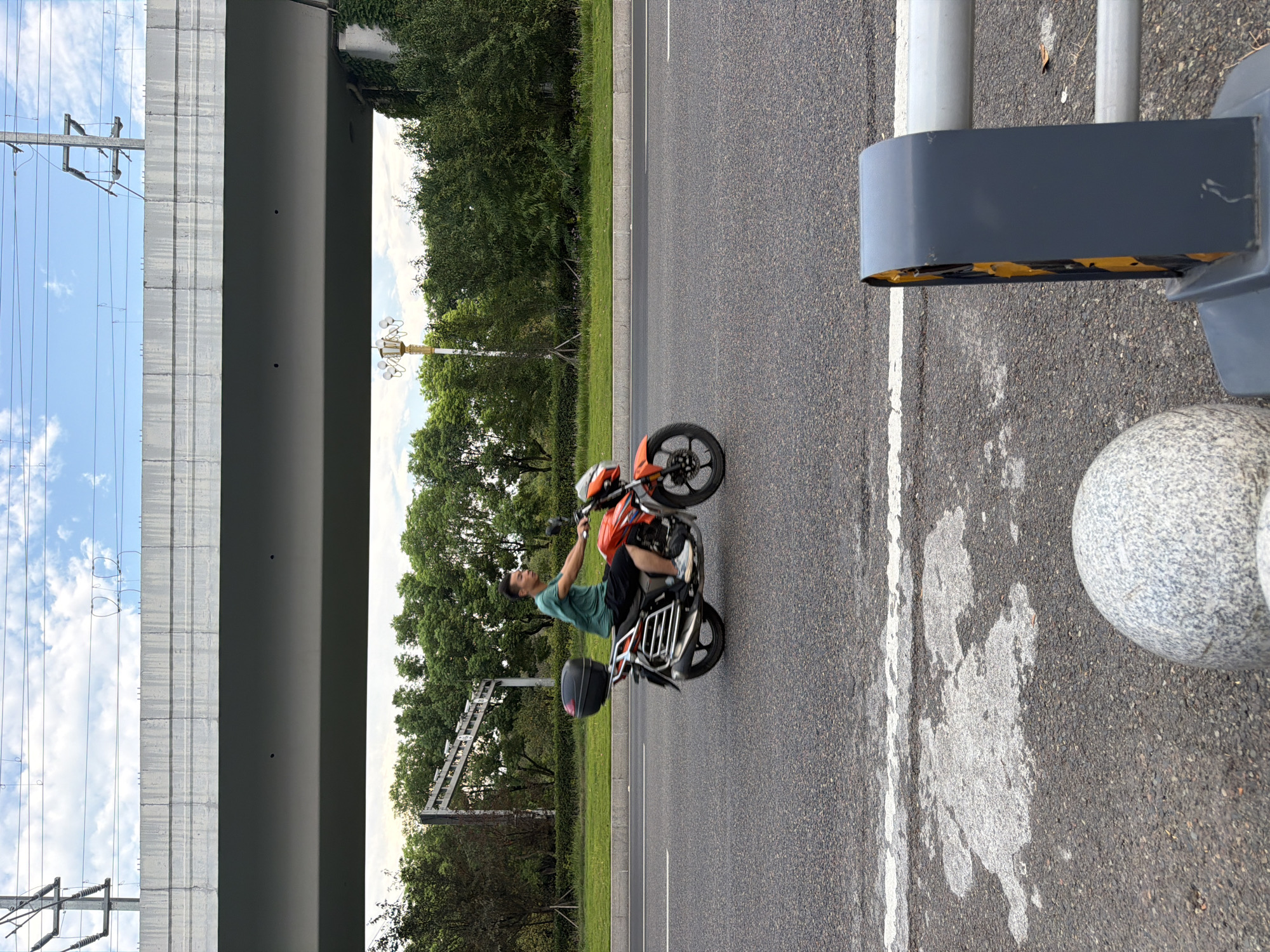 A person is riding an orange motorcycle on a paved road in Jiangjin, China. The motorcycle has a black storage box on the back and a silver rack. The rider is wearing a green t-shirt and dark shorts. The road has a white painted line. In the background, there is a concrete overpass supported by columns, with trees and greenery on either side. Overhead, there are power lines and a blue sky with scattered clouds. In the foreground, a gray decorative sphere and part of a traffic barrier are visible.