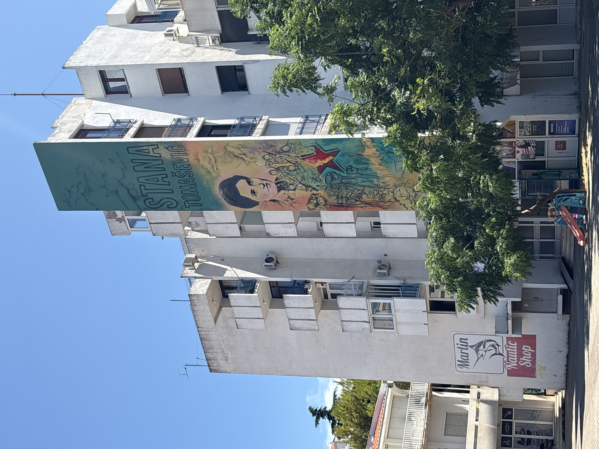A large mural of Stana Tomasevic adorns the side of a white apartment building in Bar, Montenegro. The mural features a portrait of Tomasevic with a red star and figures in the background, all rendered in vibrant colors. Below the mural, the building has several balconies and windows. At the base of the building, there are storefronts, including a "Marlin Nautic Shop" and a kiosk. Green trees partially obscure the lower portion of the building. The sky is clear and blue. In the foreground, two children are playing on a small red and blue slide. The scene appears to be on a sunny day in a residential and commercial area.