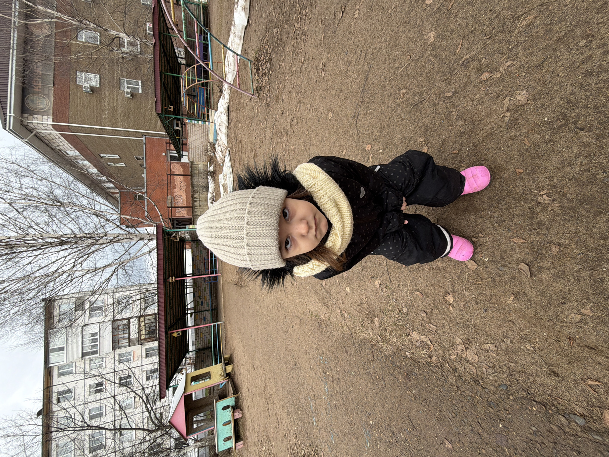 A young girl wearing a cream-colored knit hat, black polka-dot snowsuit, and pink boots stands in a playground. The playground surface is brown, possibly dirt or packed earth, with scattered leaves and debris. Behind the girl are playground structures made of metal and wood. In the background are two multi-story apartment buildings, one with a brick facade and the other white with multiple windows. Bare tree branches are visible in front of the buildings. There are remnants of snow on the ground near the playground structures. The location appears to be a residential area. Based on visual context and other available information, the likely location is Chaikovsky, Russia.