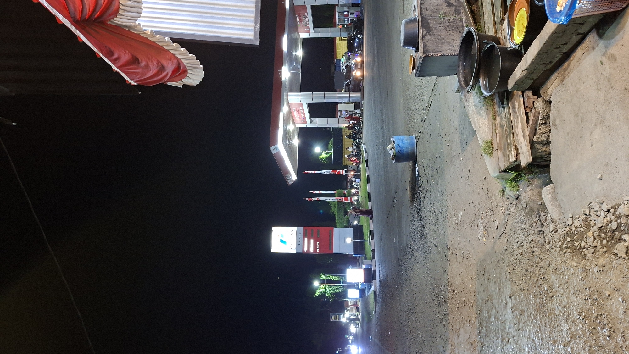 The image shows a Pertamina gas station in Keudebayu, Indonesia at night. A tall, illuminated sign displays fuel prices, with "DEXLITE" and "PERTAMAX" visible. Several motorcycles and cars are present, with some vehicles being refueled. Two flags with red and white stripes are planted in front of the station. A woman in a dark hijab and maroon dress stands on the side of the road. In the foreground, there is a blue plastic barrel and several buckets, some containing water. The ground is a mix of asphalt, gravel, and dirt. The background is dark with some distant lights. A red and white valance hangs from an awning on the right side of the frame.