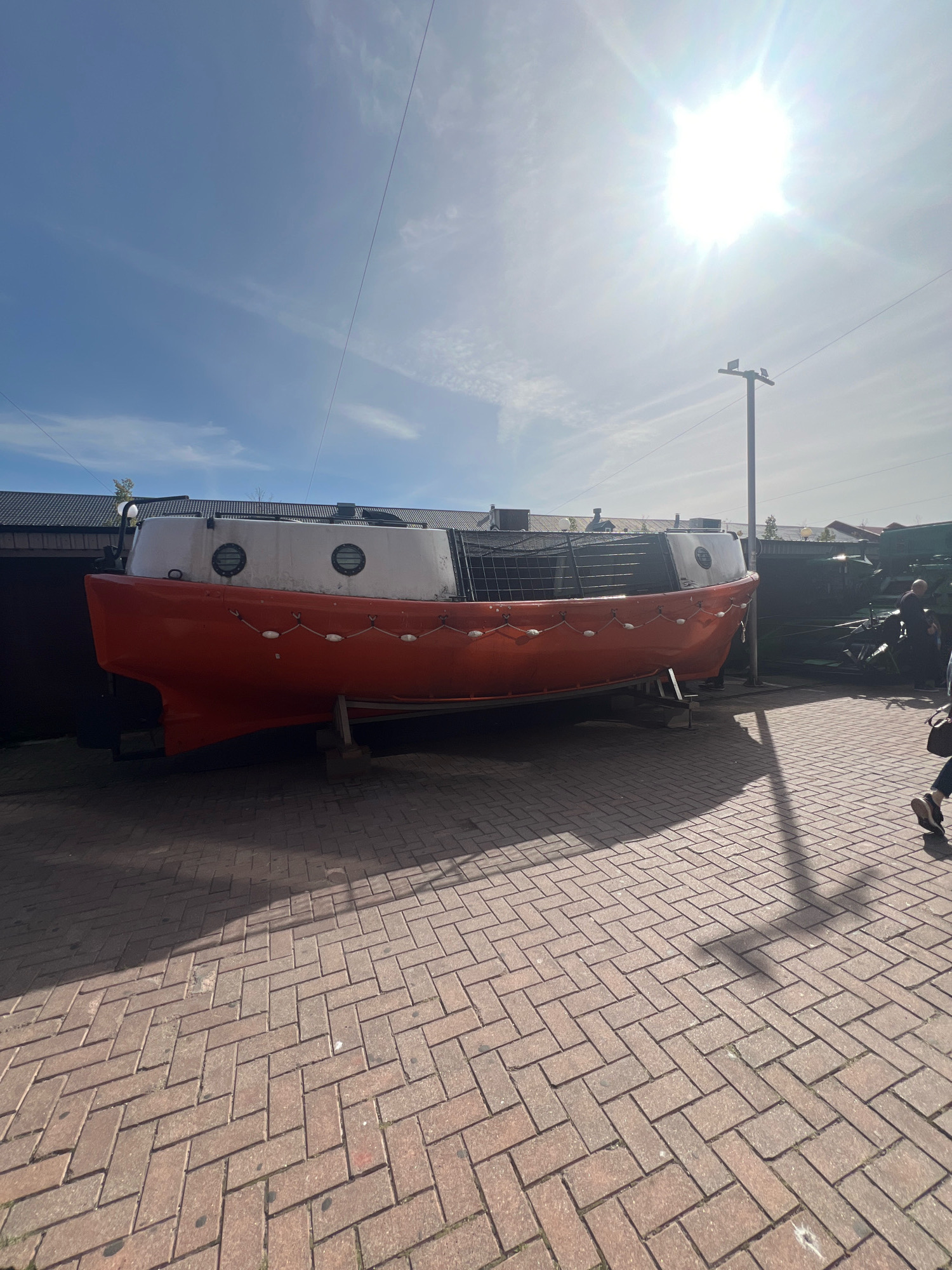 A decommissioned orange and white lifeboat rests on a brick-paved surface outside a building under a bright, sunny sky with some clouds. The lifeboat is elevated on wooden supports. A metal railing with netting is visible along the top of the lifeboat's hull. In the background, a tall pole with a light fixture stands near the lifeboat. To the right, two people are partially visible walking on the brick pavement, casting shadows. The scene appears to be an outdoor exhibition or storage area. The location is identified as the Museum of the World Ocean in Kaliningrad, Russia.