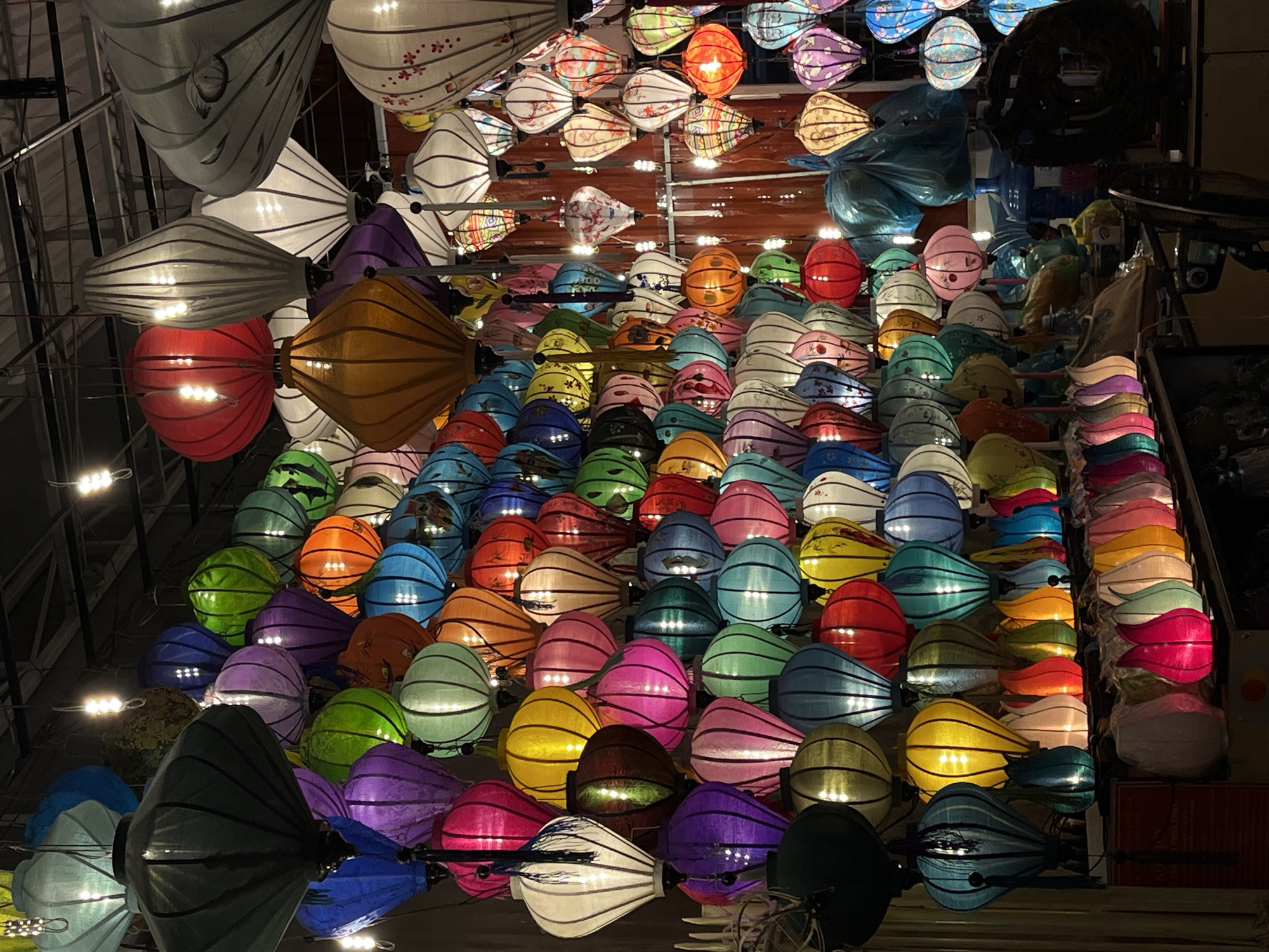 A dense arrangement of colorful illuminated lanterns occupies the frame, suggesting a location within Hội An Ward, Vietnam. The lanterns are of varying colors, including red, orange, yellow, green, blue, purple, pink, and white, with many of them being the teardrop or diamond shape associated with the city. The lamps are of different styles, with some having floral designs, others being striped, and some being solid colored. The light from the lanterns illuminates the immediate area. The lanterns appear to be arranged on shelves, or potentially suspended from the ceiling. Steel beams are visible.