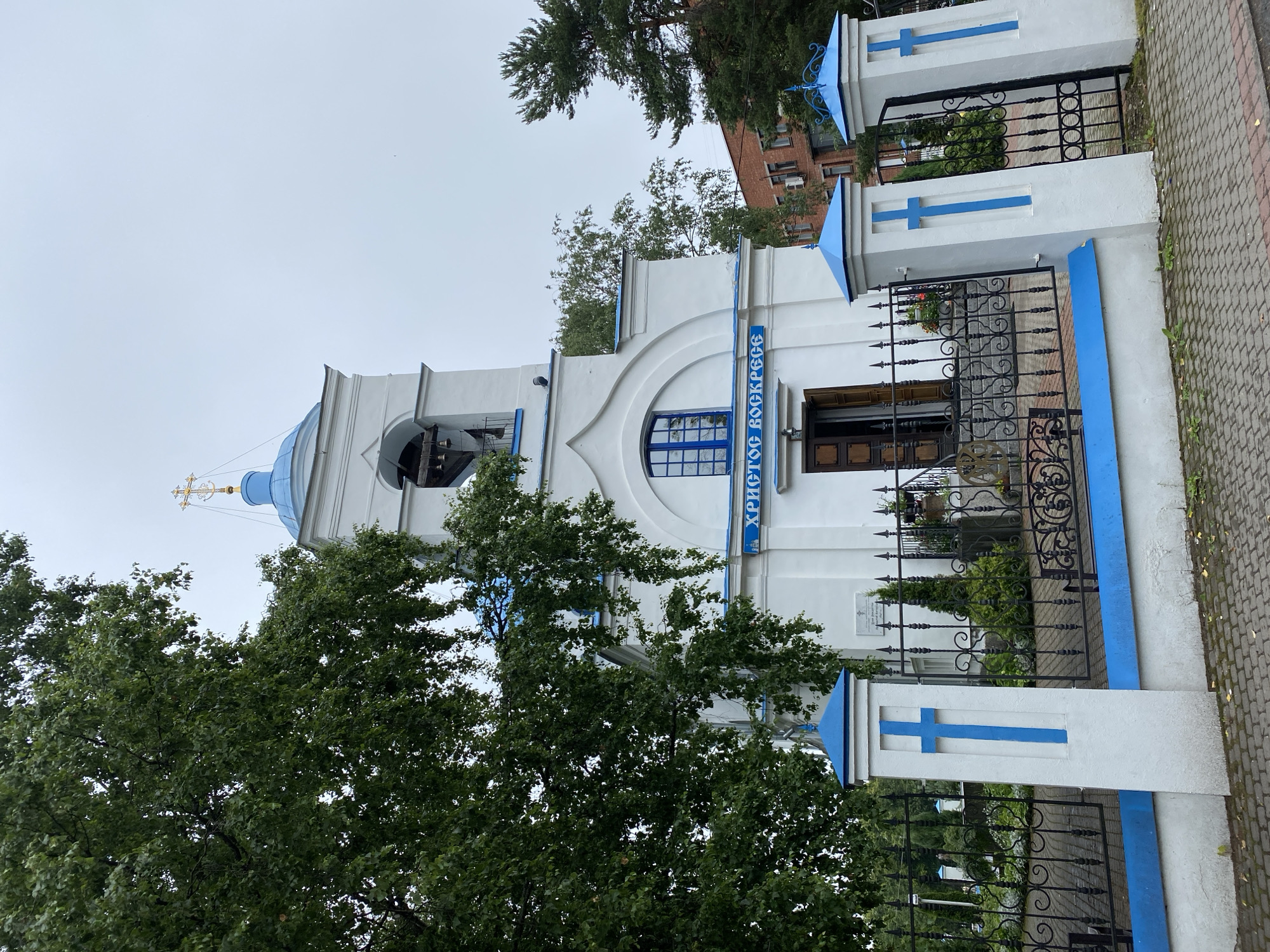 The image shows the exterior of a white Orthodox church with a blue domed bell tower topped with a golden cross. Blue accents, including crosses on the pillars and trim around the building, are prominent. The church features a blue-framed window above the entrance. The entrance is open, revealing a dark doorway. A black metal fence with decorative elements encloses the property. The grounds appear to be landscaped with greenery. In the background, a red brick building is visible. The sky is overcast. The scene is located in the city of Priozersk, Russia.