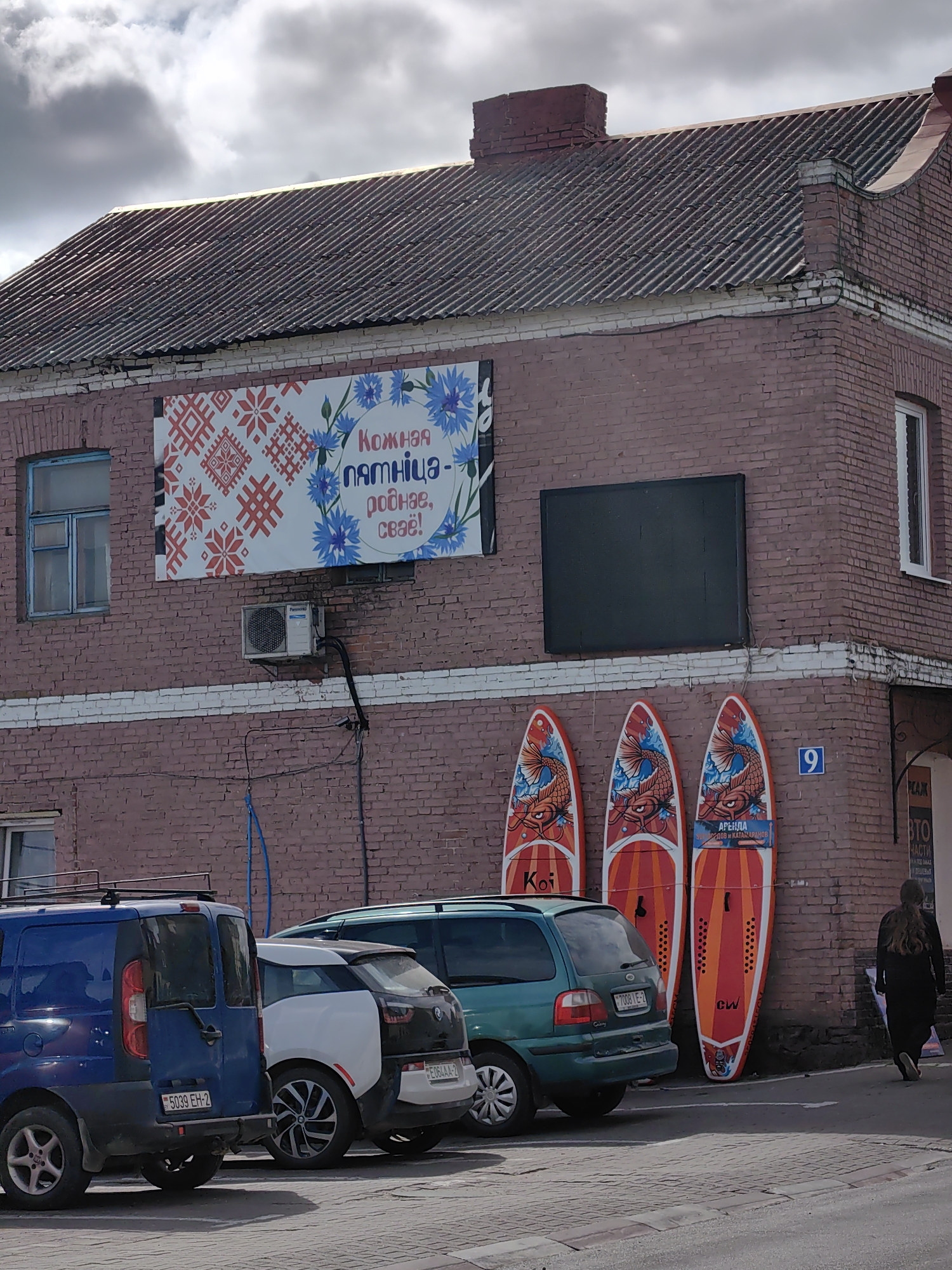 The image depicts the exterior of a building located in Pastavy, Belarus. The building features a weathered, pink brick facade and a corrugated metal roof. A large banner with red and white traditional Belarusian embroidery patterns and blue cornflowers is prominently displayed on the upper portion of the wall. Below the banner, an air conditioning unit is mounted. Three paddleboards with colorful koi fish designs are leaning against the brick wall. A blue van and a white and black BMW i3 are parked in front of the building. In the background, a green minivan and a woman with long brown hair walking away from the camera are visible. The ground is paved with interlocking bricks, and a street sign with the number '9' is attached to the wall. The sky is overcast with dramatic cloud formations.