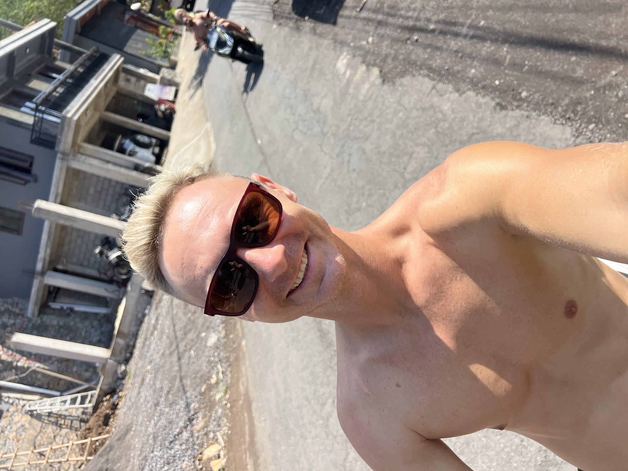 In Abang, Indonesia, a man with bleached blonde hair, wearing sunglasses, smiles at the camera while riding a scooter. He is shirtless, exposing his chest and shoulders, and appears to be in a street or road. In the background, another person on a scooter is visible, as well as buildings under construction, a parked car, and a motorcycle. The sun is shining, casting shadows on the road and ground.