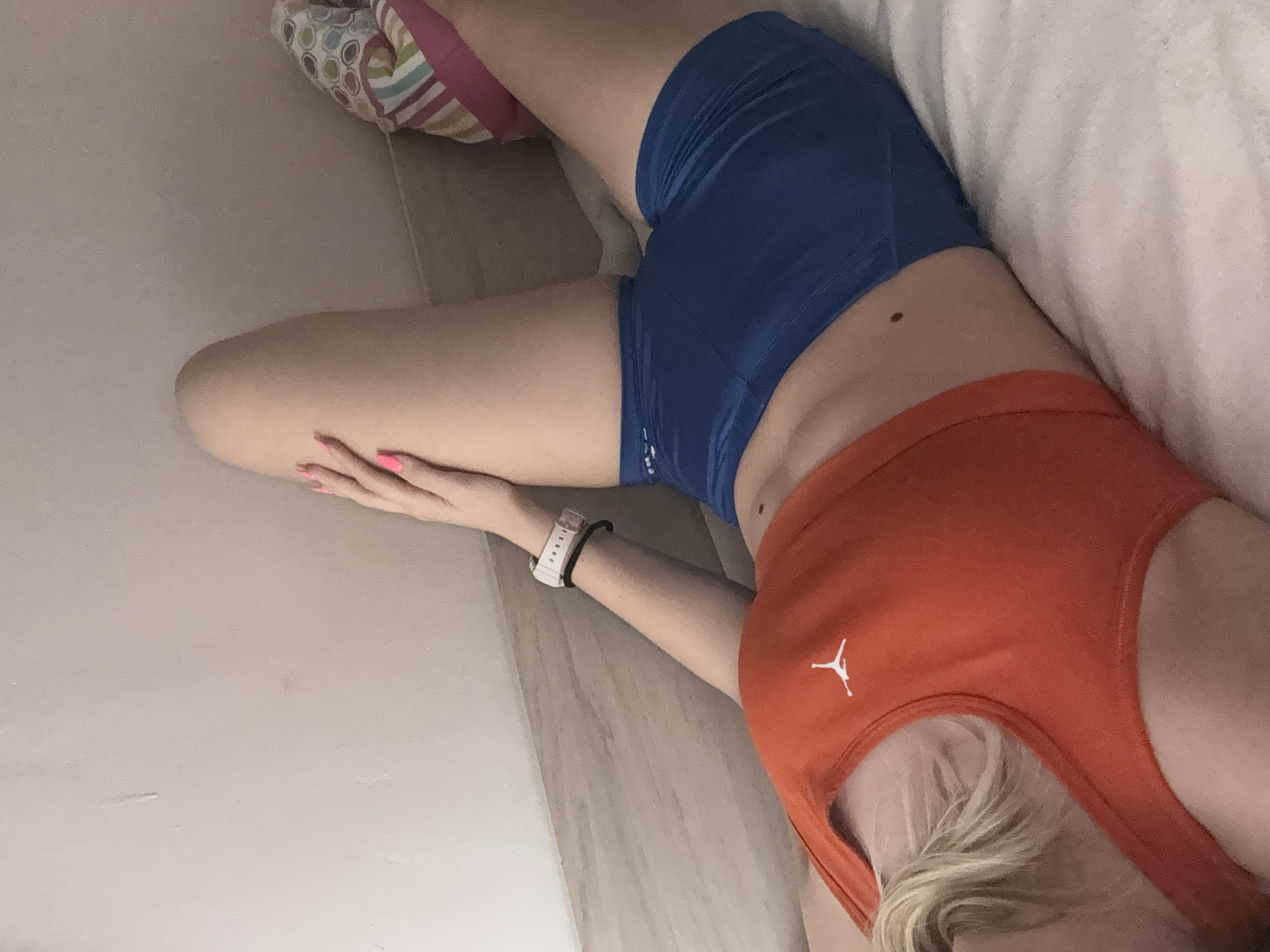 An overhead shot captures a person, likely female, lounging indoors. The individual is wearing blue athletic shorts and an orange sports bra or tank top, which features a white Jordan "Jumpman" logo on the front. A white wristwatch is visible on the left wrist, and the fingernails of the visible hand are painted bright pink. Blonde hair can be seen at the bottom right of the frame. The person is positioned diagonally, with one leg extended and a hand resting on a thigh. Several small moles are visible on the skin of the torso/hip area. The background includes a light-colored floor or wall, a white pillow or bedding material, and a multi-patterned cushion or pillow near the upper left corner of the frame. The setting suggests a relaxed, indoor environment, possibly in Sofia, Bulgaria.