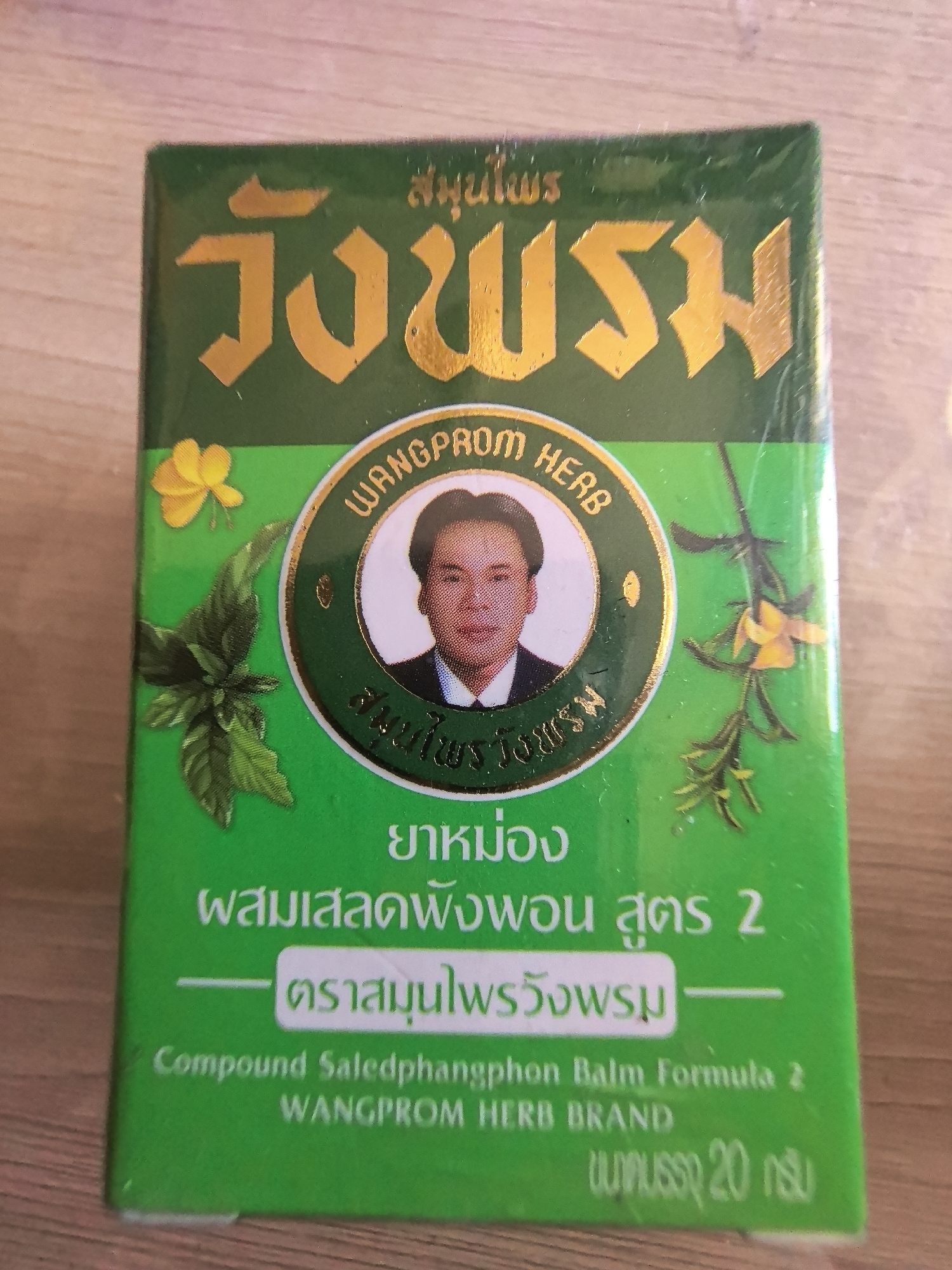 The image shows a green box of "Wangprom Herb" compound Saledphangphon balm formula 2, a Thai herbal remedy. The box features gold lettering in Thai and English, a central circular logo containing a portrait of a man, and illustrations of yellow flowers and green foliage. The text also indicates that it is a "Wangprom Herb Brand" and that the net weight is 20 grams. The background is a wooden surface.