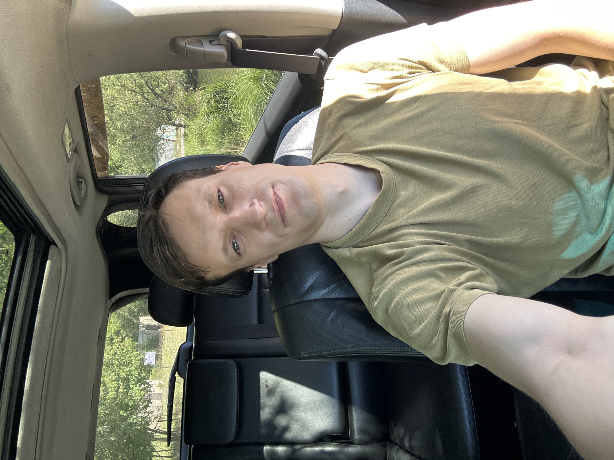 A man with short brown hair is sitting in the driver's seat of a car. He is wearing a tan t-shirt. The interior of the car is visible, including black leather seats. The sunroof is open, and sunlight is streaming in. Outside the car, there are trees and green grass. The scene appears to be in a rural or suburban area, possibly on the outskirts of Makiv, Ukraine, given the context provided. The man is looking at the camera, and his expression is neutral. He is holding his right arm out of frame, likely taking the picture.