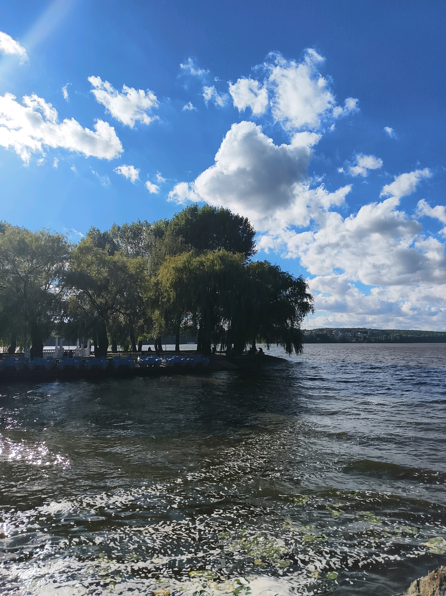 The image captures a scenic view of the Ternopil Pond in Ternopil, Ukraine, on a bright, partly cloudy day. In the foreground, the water is dark and rippled, with patches of white foam and greenery floating on the surface. The sun glints off the water, creating a shimmering effect.
Along the shore, a line of lush green trees forms a dense canopy, with their branches extending towards the water. Some of these appear to be weeping willow trees, characterized by their long, drooping branches. The trees create a natural barrier between the water and a paved area behind them, where small paddle boats are lined up, some with covers. There are a few indistinct figures visible under the trees.
The background features a wide expanse of water, stretching towards a distant, tree-covered shoreline. A faint line of buildings or structures can be seen on the horizon, suggesting the presence of a town or city. The sky above is a vibrant blue, dotted with fluffy white clouds. The sun is positioned high in the sky, casting rays that create a bright spot and lens flare on the left side of the image. The overall scene depicts a tranquil and picturesque waterfront setting.