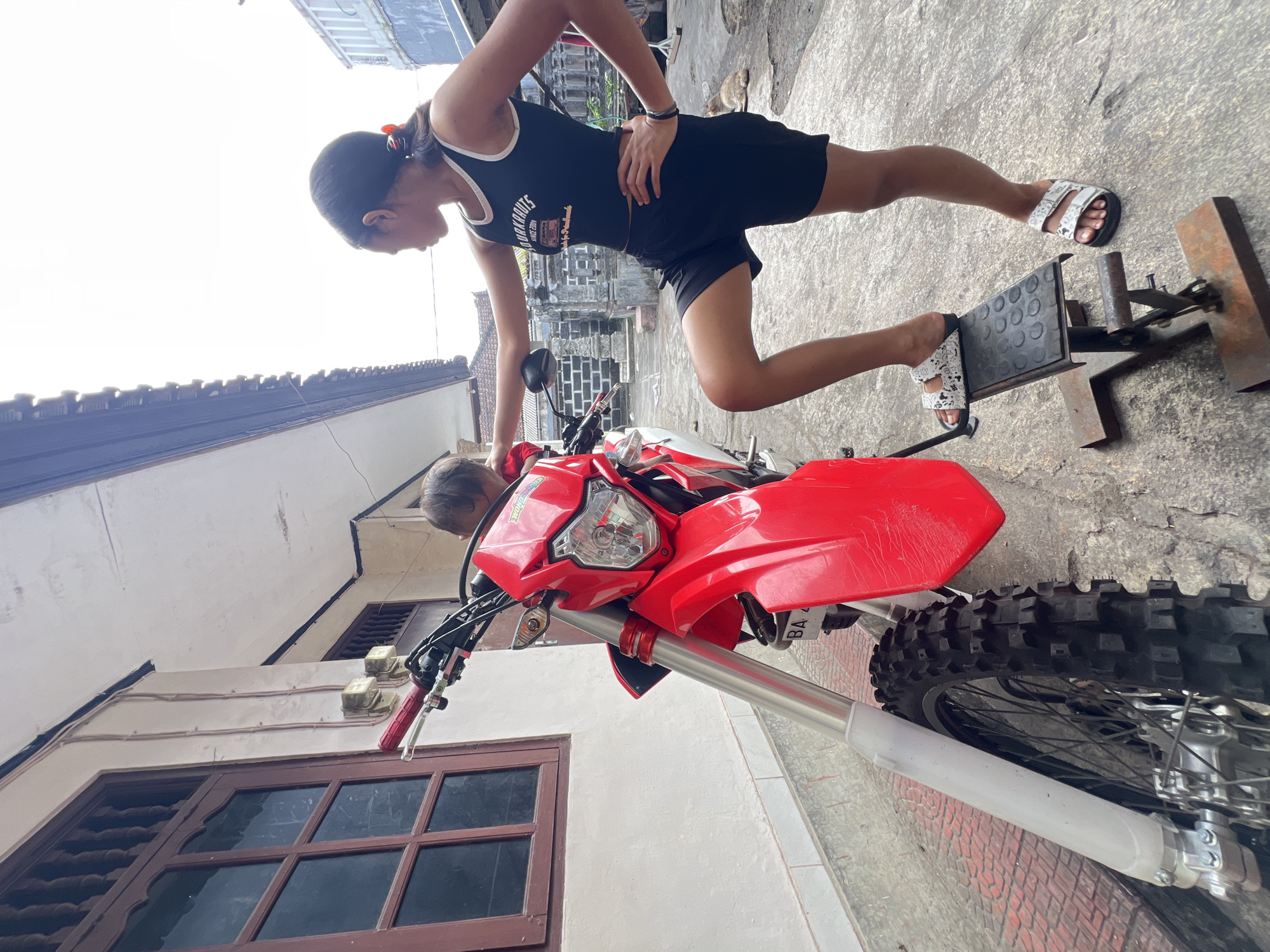 A medium shot captures a young woman standing beside a red dirt bike on a paved outdoor area, likely a residential alley or driveway, in Denpasar, Indonesia. The woman, wearing a black tank top and shorts, has her left hand on the dirt bike and her right hand on her hip. Her left leg is bent and resting on a metal stand positioned next to the bike. She is looking towards the left of the frame. The dirt bike is predominantly red with silver accents on its front suspension. To the left of the bike is a building with a light-colored wall and a wooden-framed window with dark panes. The sky is overcast. In the background, there are more buildings and what appears to be a glimpse of some traditional Balinese architecture.