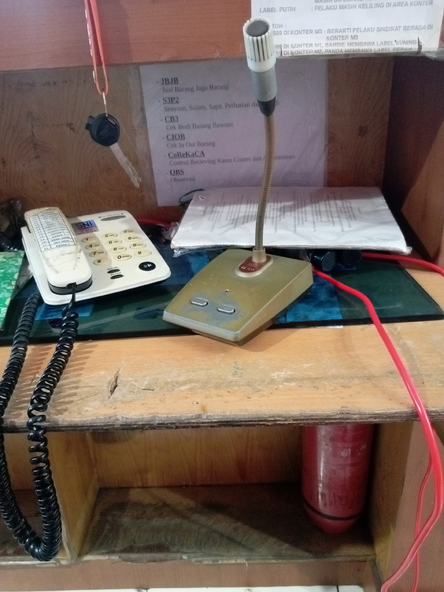 The image depicts a shelf in what appears to be a security or service counter in Batam, Indonesia. On the shelf are various objects including a white corded telephone with a keypad and a display screen, a gooseneck microphone with a tan base, a stack of papers, and a red coiled cord. A Honda key is hanging from a pink lanyard on the left side of the frame. In the background, a sign lists various codes and descriptions such as "JBJB - Jual Barang Jaga Barang", "S3P2 - Senyum, Salam, Sapa, Perhatian dan...", "CB3 - Cek Bodi Barang Bawaan", "CIOB - Cek In Out Barang", "CoReKaCA - Control Recieving Kassa Counter dan Administrasi", and "OBS - Observasi". Another section of the sign has text referring to "LABEL PUTIH" and "S09 DI KONTER M3". The shelf is made of worn wood, and below it, a red cylindrical object, possibly a fire extinguisher or a water bottle, is visible in a recessed compartment. The overall environment suggests a functional, possibly dated, operational space.