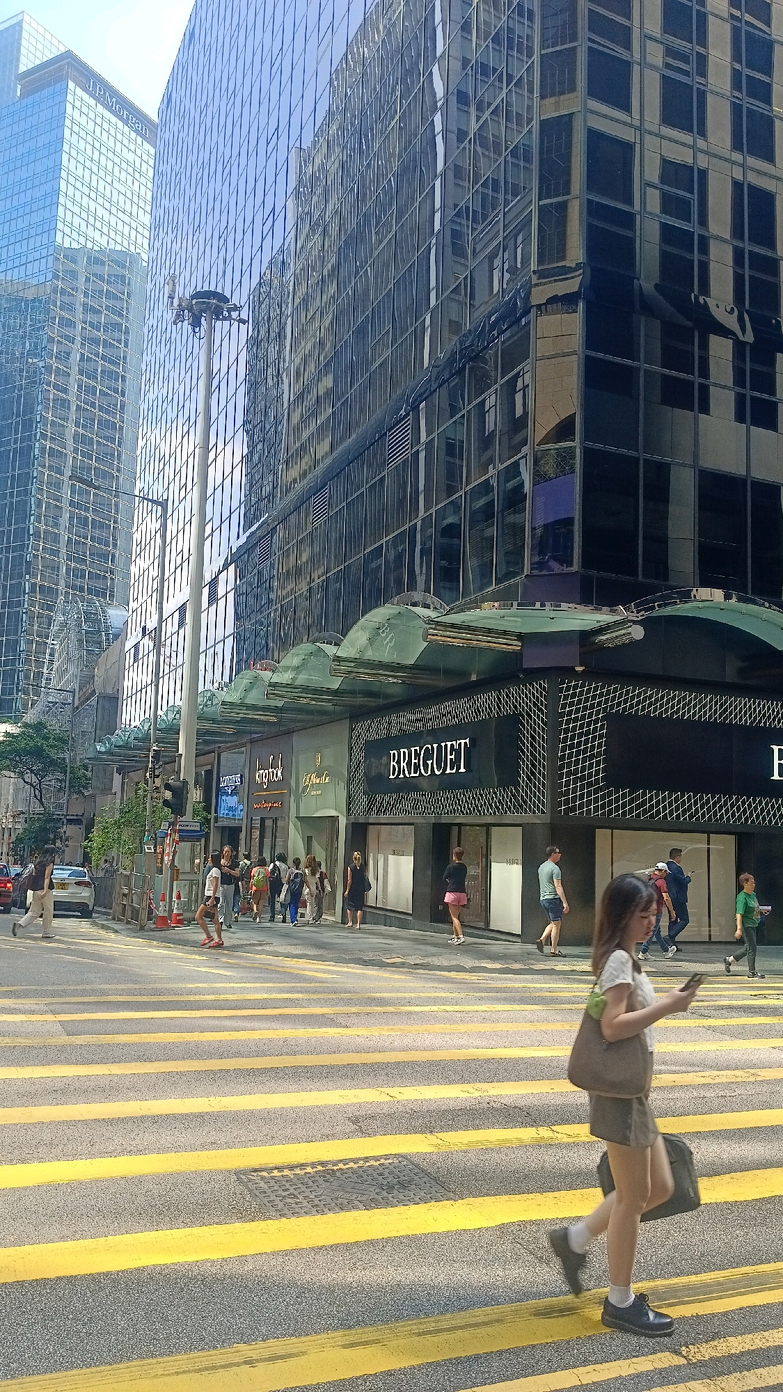The image depicts a street scene in Hong Kong, China, likely in a commercial district characterized by modern high-rise buildings with reflective glass facades. The foreground features a prominent crosswalk with thick yellow stripes. Several pedestrians are visible, crossing the street or walking on the sidewalk.
In the middle ground, a woman in a white top and brown skirt is crossing the street, holding a smartphone in her right hand and carrying a grey tote bag over her left shoulder. Another person is seen walking behind her, carrying a black bag. On the sidewalk, a group of people are walking, and the storefronts of several luxury brands, including "BREGUET," "Kingbook," and "J. Laumer & Cie," are visible.
The background is dominated by large, glass-fronted buildings, reflecting the sky and other structures. One building prominently displays the "J.P. Morgan" logo. A tall lamppost with a light fixture stands near the sidewalk. The sky is clear and bright, suggesting daylight. The overall impression is a busy urban environment with a focus on retail and finance.