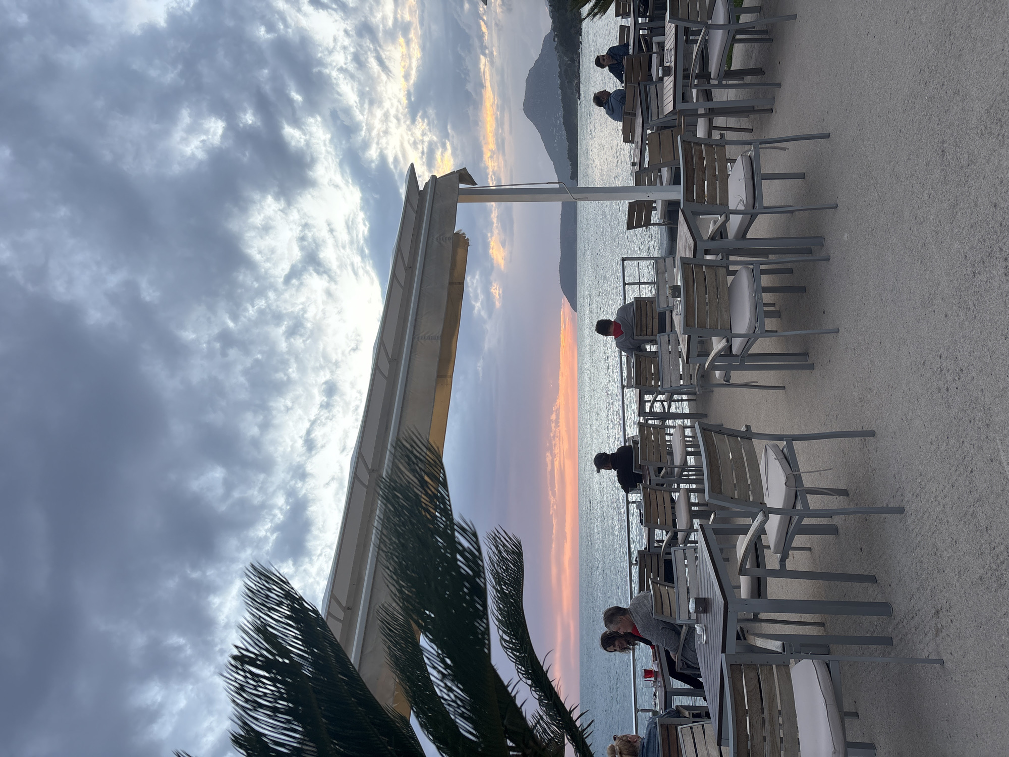 The image shows a sunset view from a seaside restaurant in Bar, Montenegro. Several people are seated at tables, likely enjoying the view. The tables and chairs are a modern, gray metal and wood design. A partial awning is visible, along with a palm tree. The sky is cloudy with a vibrant sunset glow on the water. A mountain is faintly visible in the background across the bay.