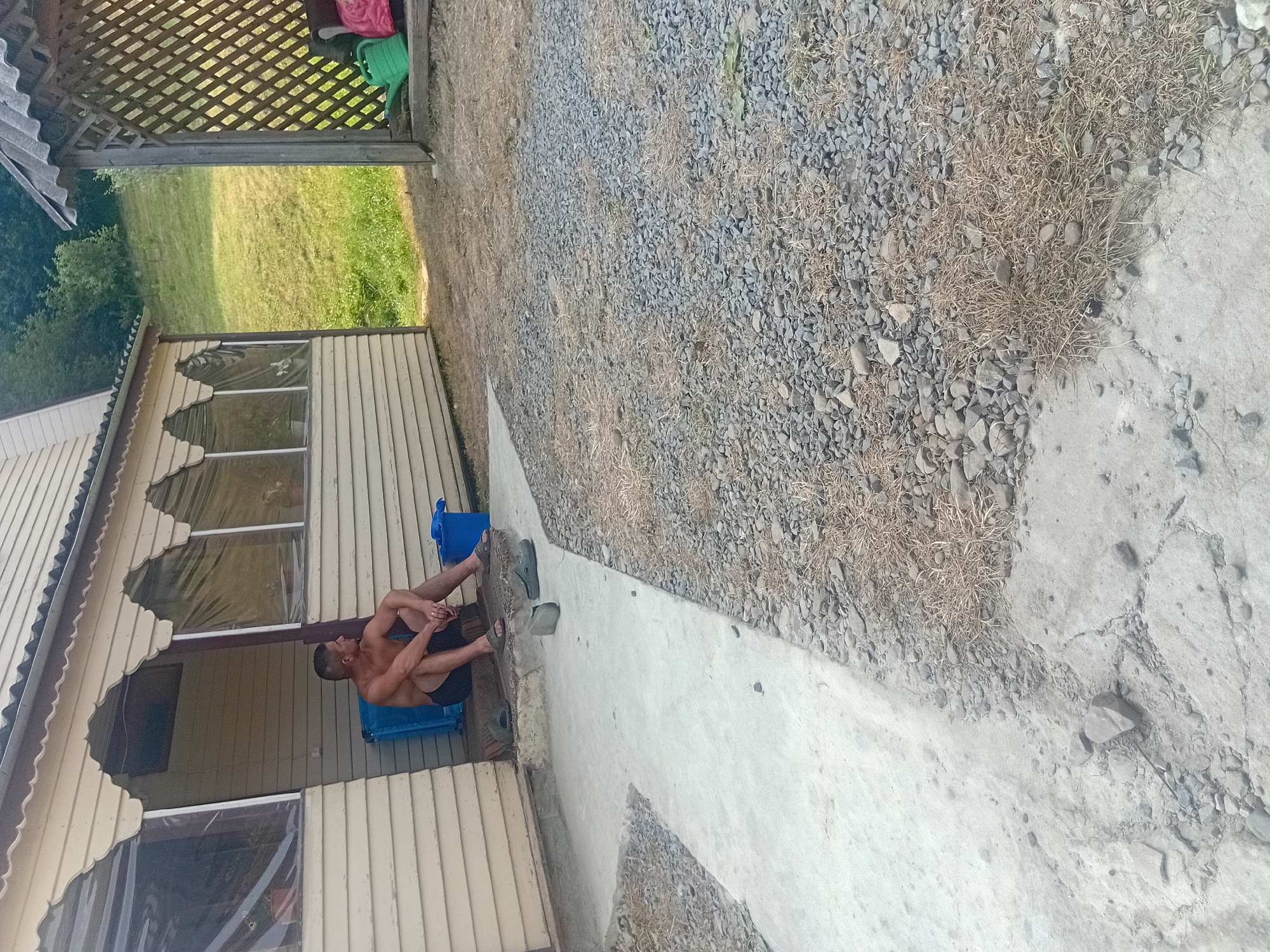 The image depicts an outdoor scene on what appears to be a sunny day. A man is sitting on a blue plastic chair on a covered porch next to a light-colored wooden building. He is wearing black shorts and is shirtless. The porch has a wooden railing and windows covered with clear plastic. The building itself has a light yellow exterior.
Beyond the porch, the ground is covered with gravel and dry grass. A concrete pathway leads away from the porch. In the background, a green lawn can be seen, suggesting a rural or semi-rural setting. There is also a wooden lattice structure and a green watering can and a pink object visible on the porch area.
The man appears relaxed, with his arms resting on his knees. The overall atmosphere is casual and likely indicative of a warm day, possibly during summer given the attire and outdoor setting. No text is visible in the image. The location context is Verkhniachka, Ukraine.