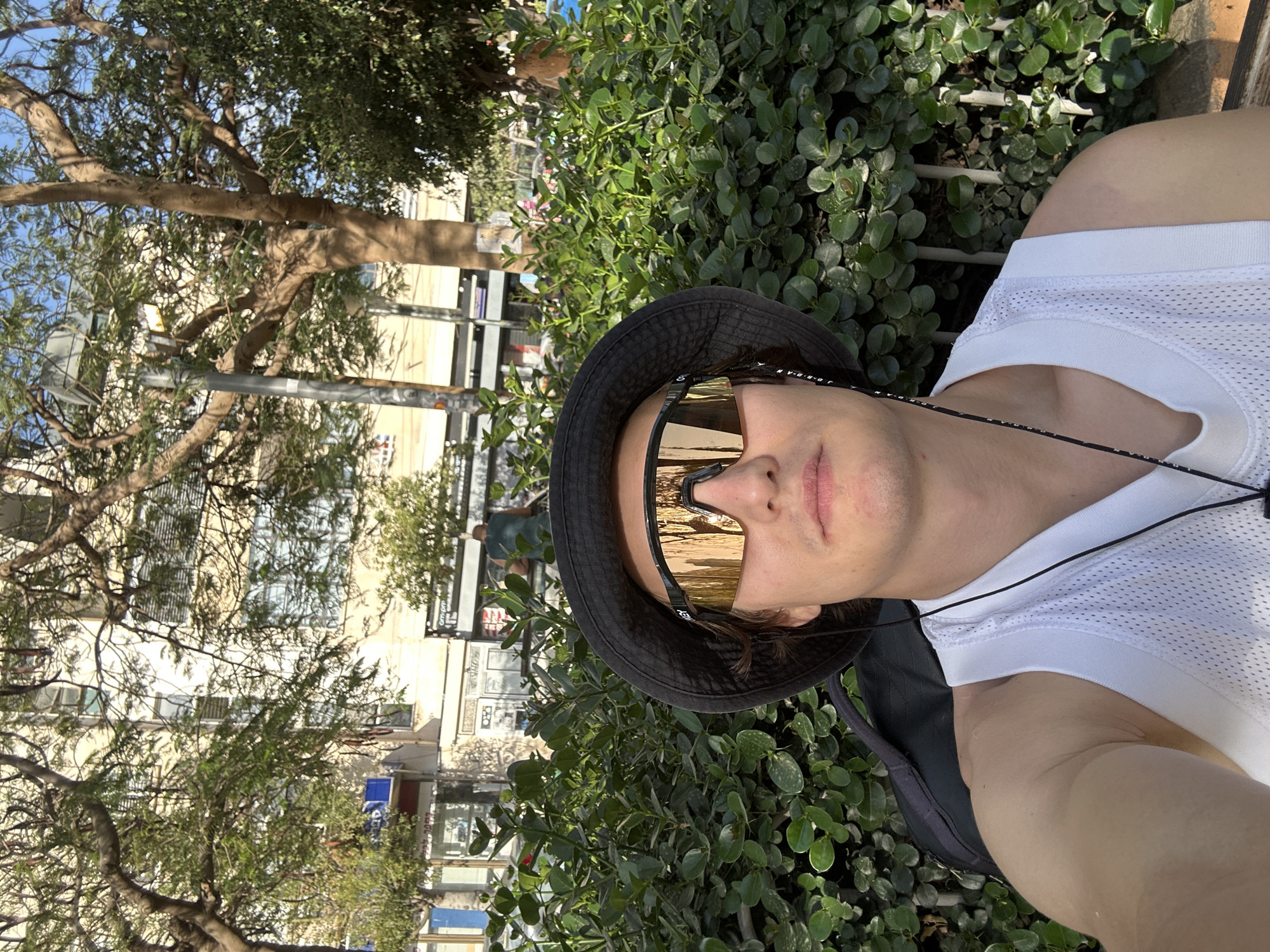 A selfie shows a person with light skin, wearing gold reflective sunglasses and a black bucket hat, and a white mesh tank top. The person is positioned in front of a dense green shrub. In the background, blurred buildings and trees suggest an urban environment. The buildings are light-colored, and there is a glimpse of storefronts with signage. The presence of Hebrew lettering on signs suggests the location is likely Tel Aviv, Israel.