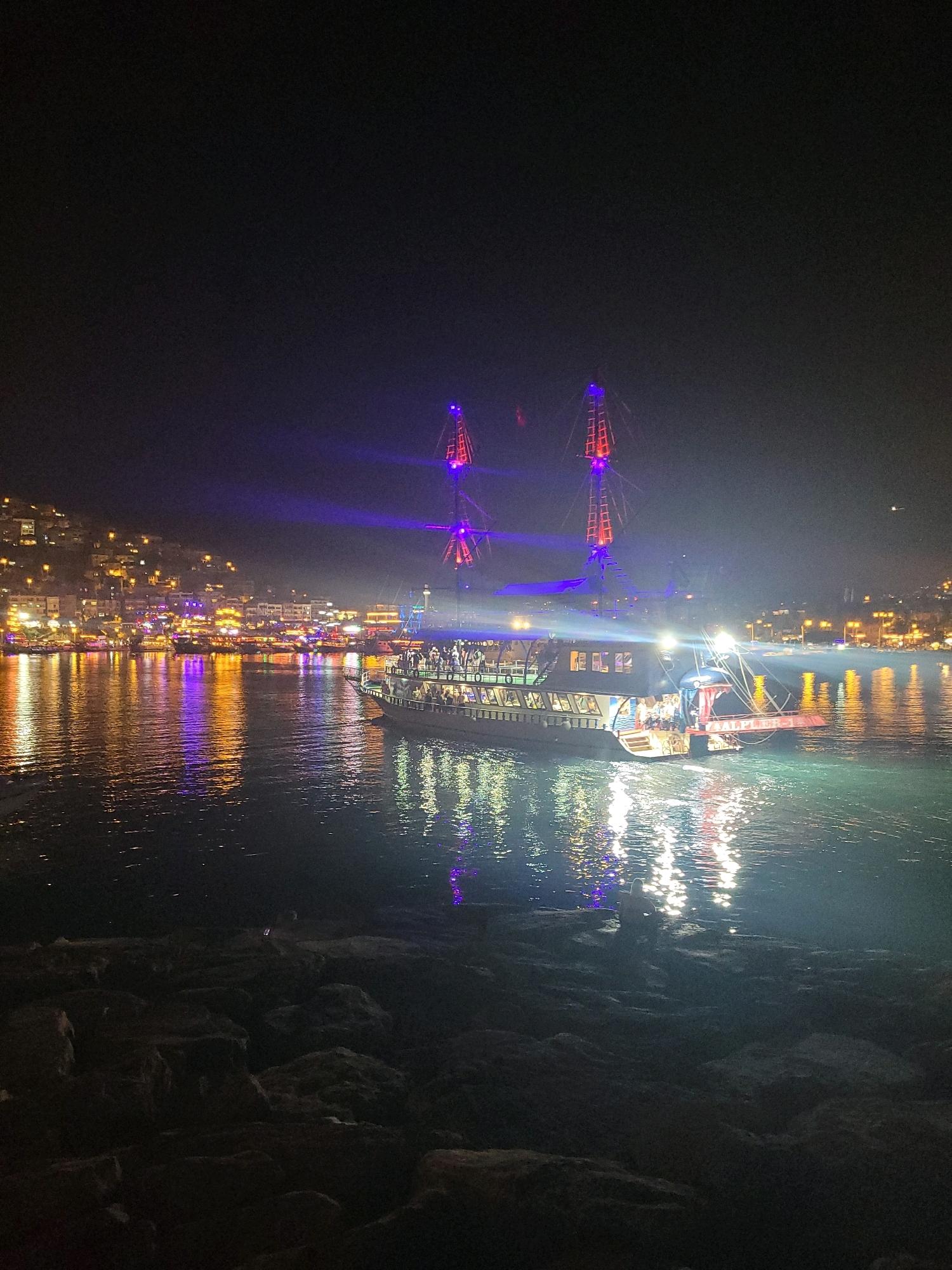 The image depicts a nighttime scene in Alanya, Turkey, featuring a pleasure boat with illuminated masts on the water. The boat is prominently positioned in the foreground, with its decks visibly populated by people enjoying the evening. Searchlights from the boat cast colorful beams of purple and red light into the dark sky. In the background, the Alanya coastline is illuminated with numerous lights from buildings, indicating a densely populated urban area. The water reflects the vibrant lights from the boat and the city, creating a dynamic visual effect. The foreground is occupied by dark, rocky terrain, possibly a harbor wall or shoreline.