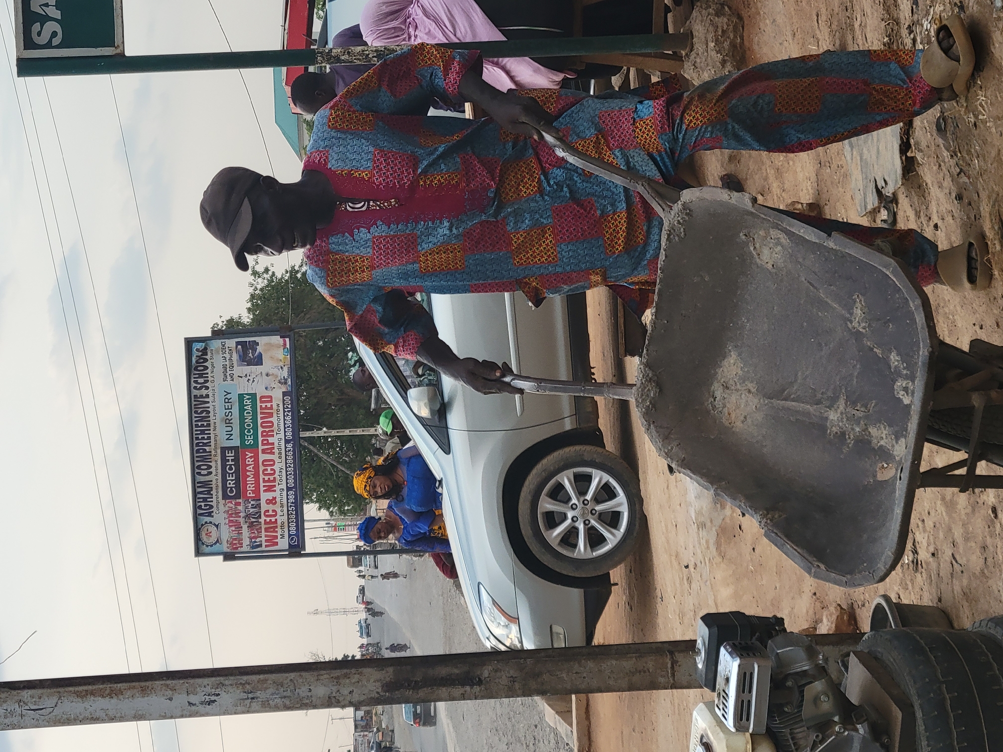 The image shows a man in patterned clothing operating a wheelbarrow. He is on a dirt surface next to a parked white car with people inside. In the background, there is a street with traffic and a sign that reads "AGHAM COMPREHENSIVE SCHOOLS" which indicates this could be near a school in Abuja, Nigeria.