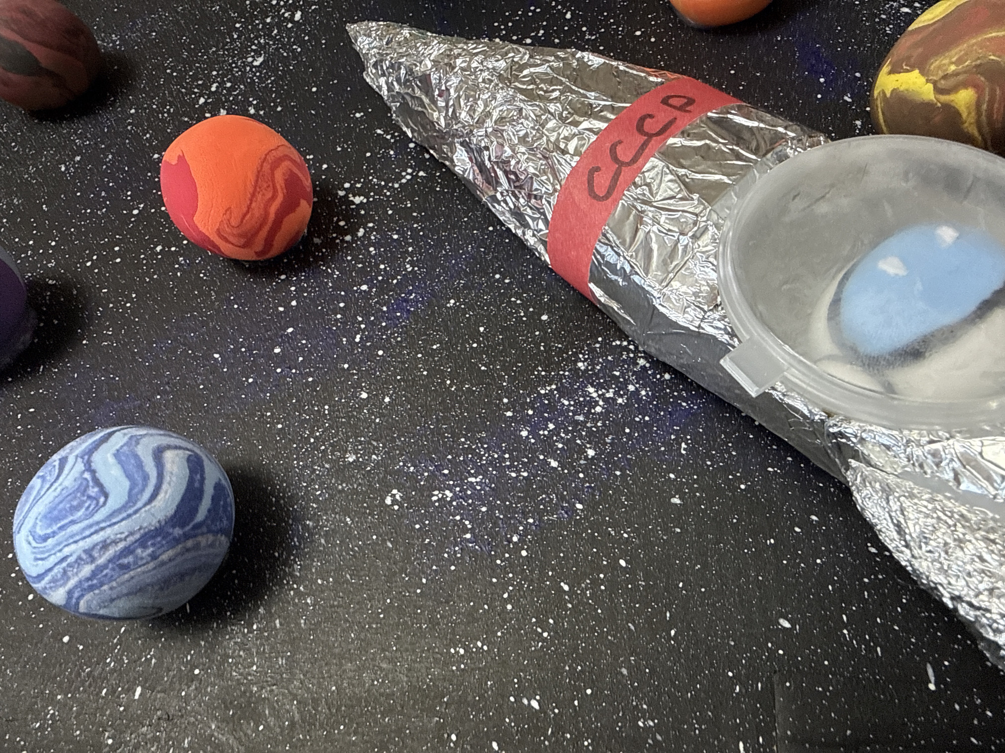 A tabletop diorama depicts a space scene on a black surface speckled with white dots, simulating stars. Scattered around are various spherical objects resembling planets, each with unique color patterns. A rocket made from aluminum foil with a red band labeled "CCCP" lies at an angle to the upper right. The back of the rocket has a clear plastic cup affixed to it, containing a small, light blue, spherical object. The location within the city of Chaikovsky, Russia cannot be determined from the image. There are no people present in the image.
