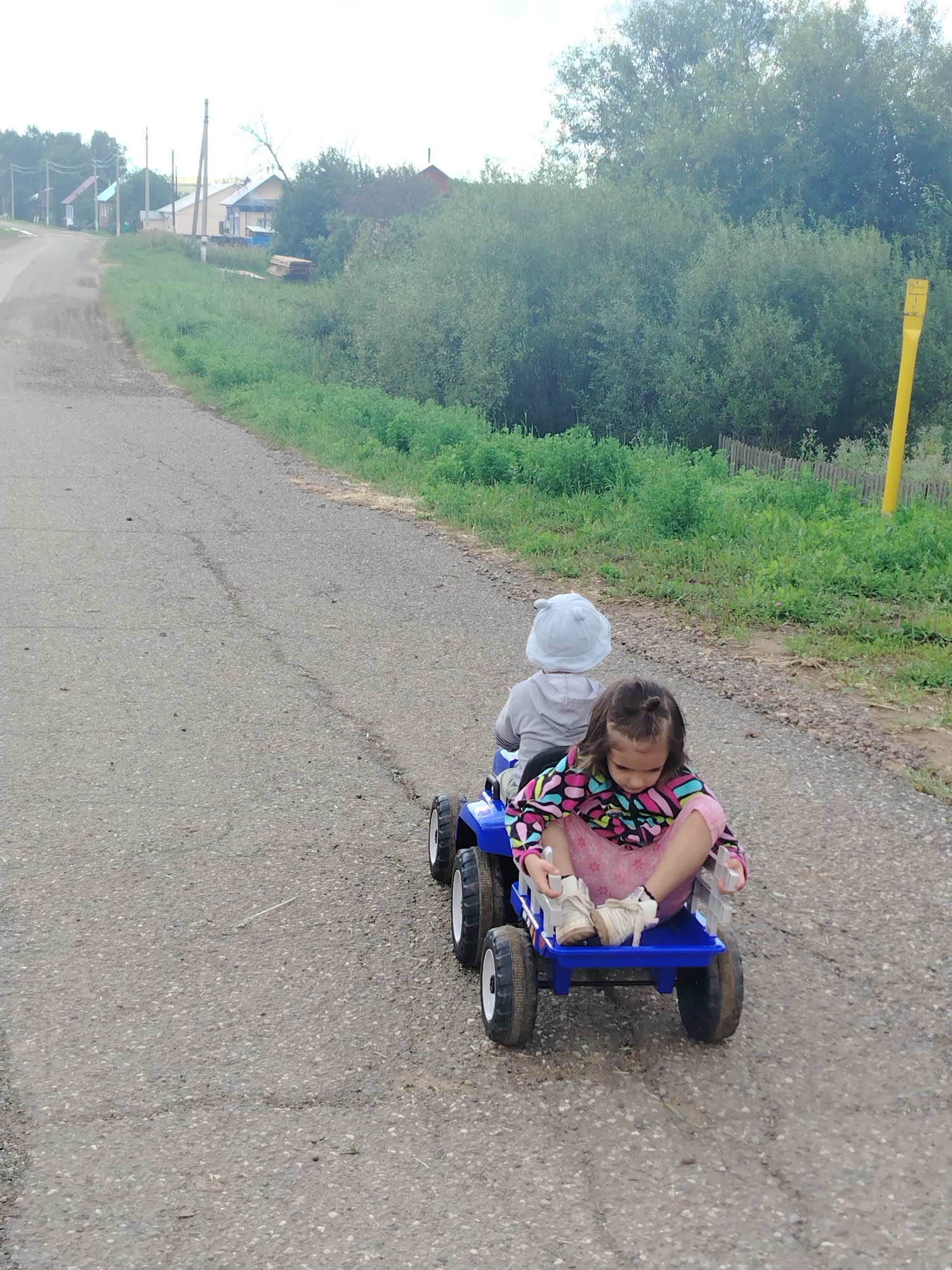 Two young children are riding in a blue toy tractor on a paved road. The child in front, a girl, is wearing a pink and black patterned shirt, pink pants with stars, and white shoes. She is sitting facing forward. The child behind her, a boy, is wearing a grey hooded sweatshirt and a light grey hat. He is facing away from the camera. The tractor is blue with black wheels and white rims. The road is asphalt and appears to be in a rural area, with a grassy verge on the right side of the road. In the background, there are trees and some houses, suggesting a village or small town setting. A yellow post is visible on the right side of the road, and a wooden fence can be seen further back. The overall environment appears to be outdoors on a cloudy day.