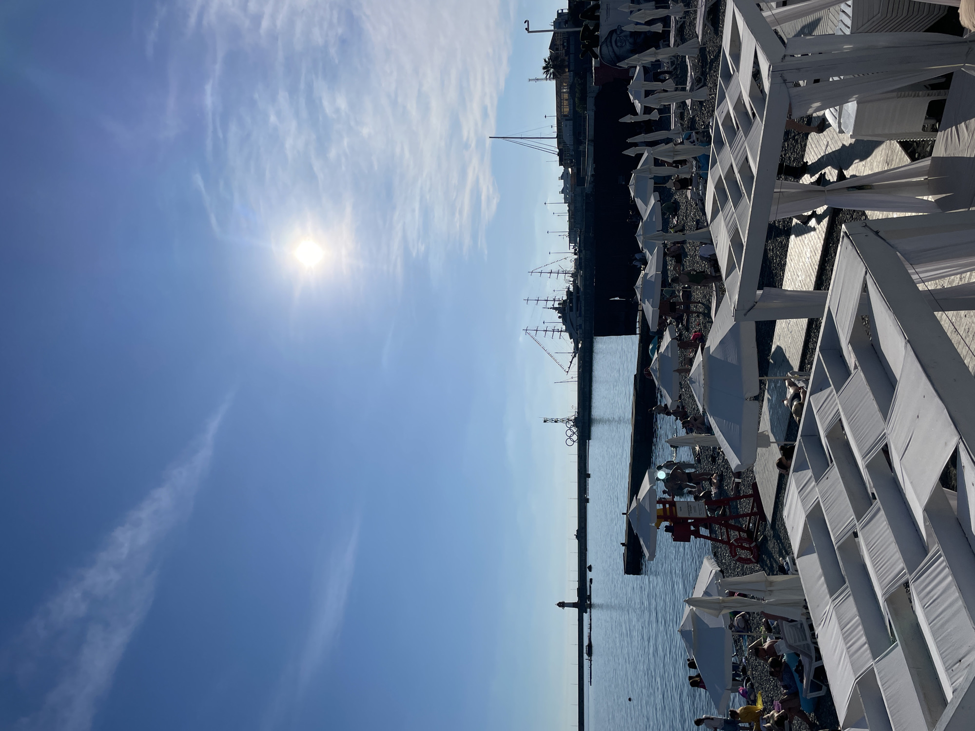 The image depicts a sunny day at a pebble beach in Sochi, Russia, likely near the port. In the foreground, white canopy beds with white drapes are set up on wooden planks. Beyond them, white beach umbrellas are scattered across the pebble beach, with several people relaxing on lounge chairs and towels. A red lifeguard stand is visible with a person sitting on it. The sea stretches out to the horizon, where a lighthouse stands on a pier to the left, and a harbor with several docked boats, including a large sailing ship and a yacht, is situated in the middle. The sky is blue with scattered wispy clouds, and the sun is bright, casting a glare. The Olympic rings symbol is visible on a structure near the harbor. The overall scene is one of leisure and a popular tourist destination during daylight hours.
