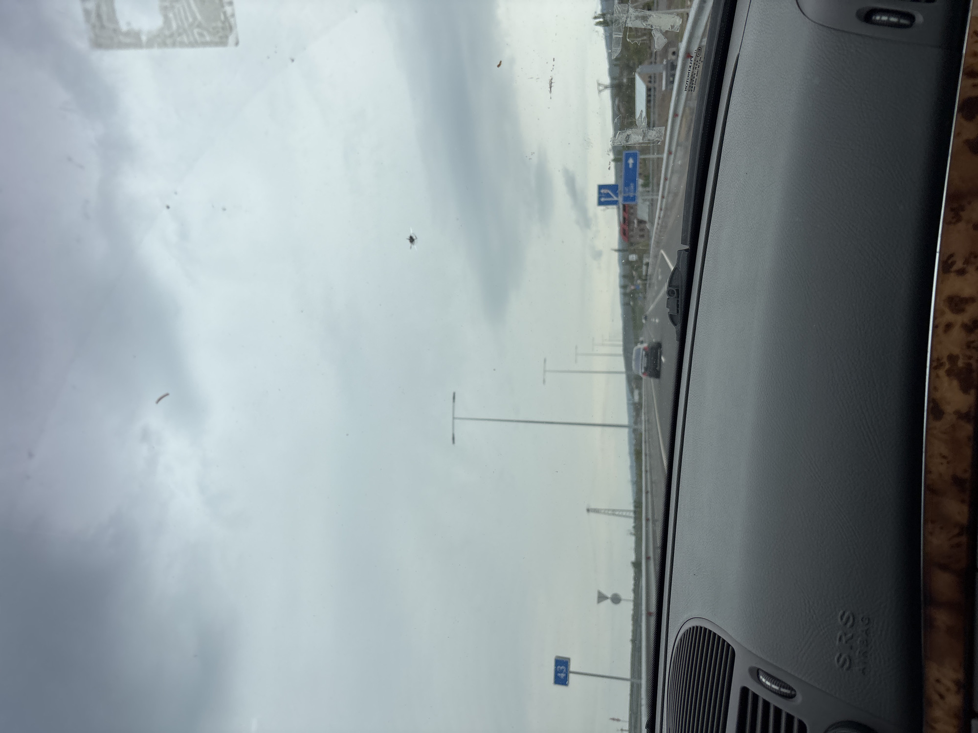 The image shows a view from inside a car, looking out through the windshield. It appears the car is driving on a highway or major road. On the side of the road, there are lampposts and road signs. The sky is overcast. The dashboard of the car is visible in the foreground.
It is difficult to say for sure, but the location is not in the city of Kosh, Armenia.