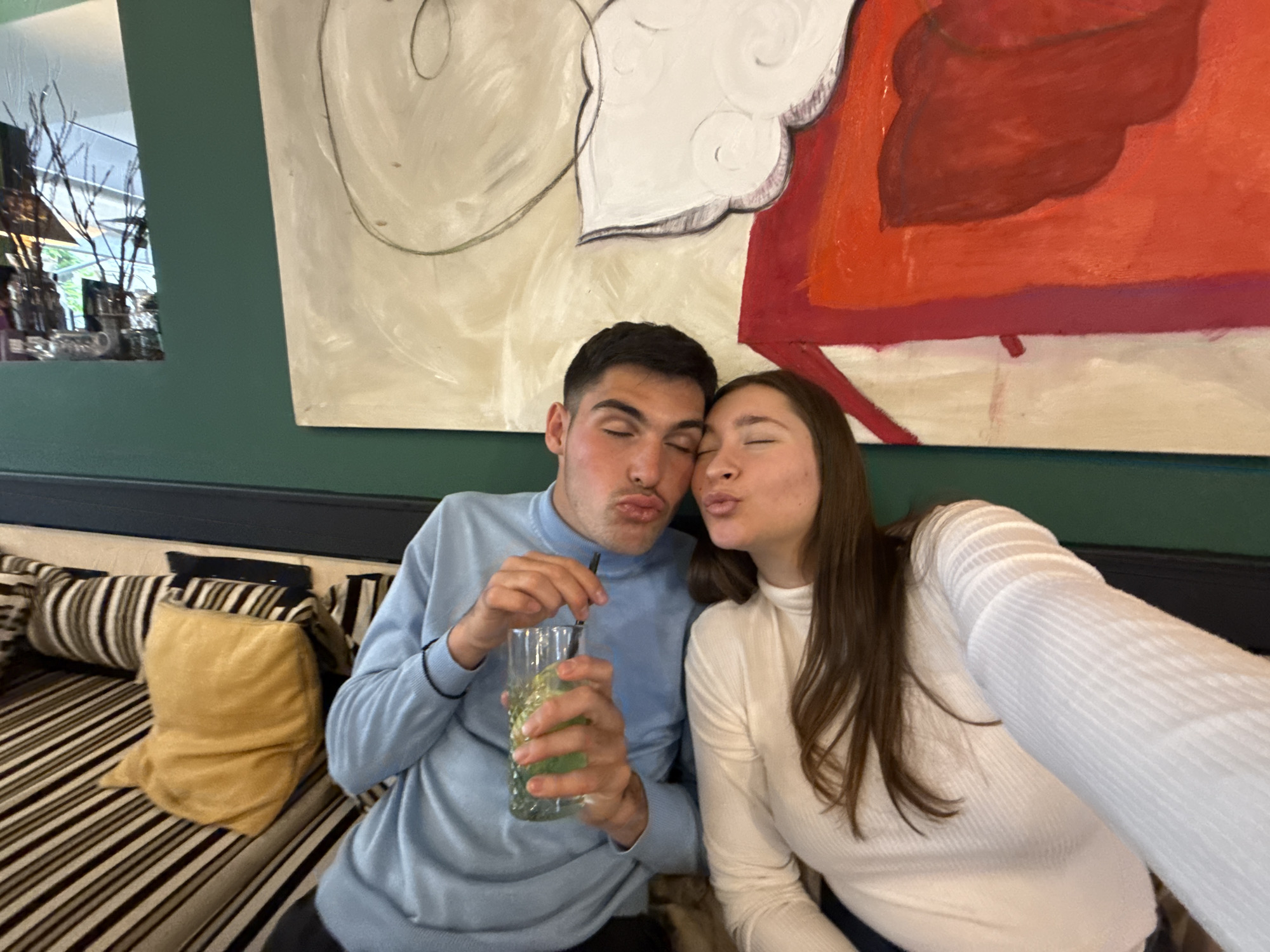 Two people, a male and a female, are sitting together on a striped couch. The male is wearing a blue sweater and holding a glass with a green liquid and a straw. The female is wearing a white top and is holding up the cell phone taking the picture. They both appear to be making a kissing face towards the camera. Behind them is a green wall with a large painting. There is also a partial view of a window reflection in the upper left corner. It is not possible to definitively identify the location as Sofia, Bulgaria from the given visual information.