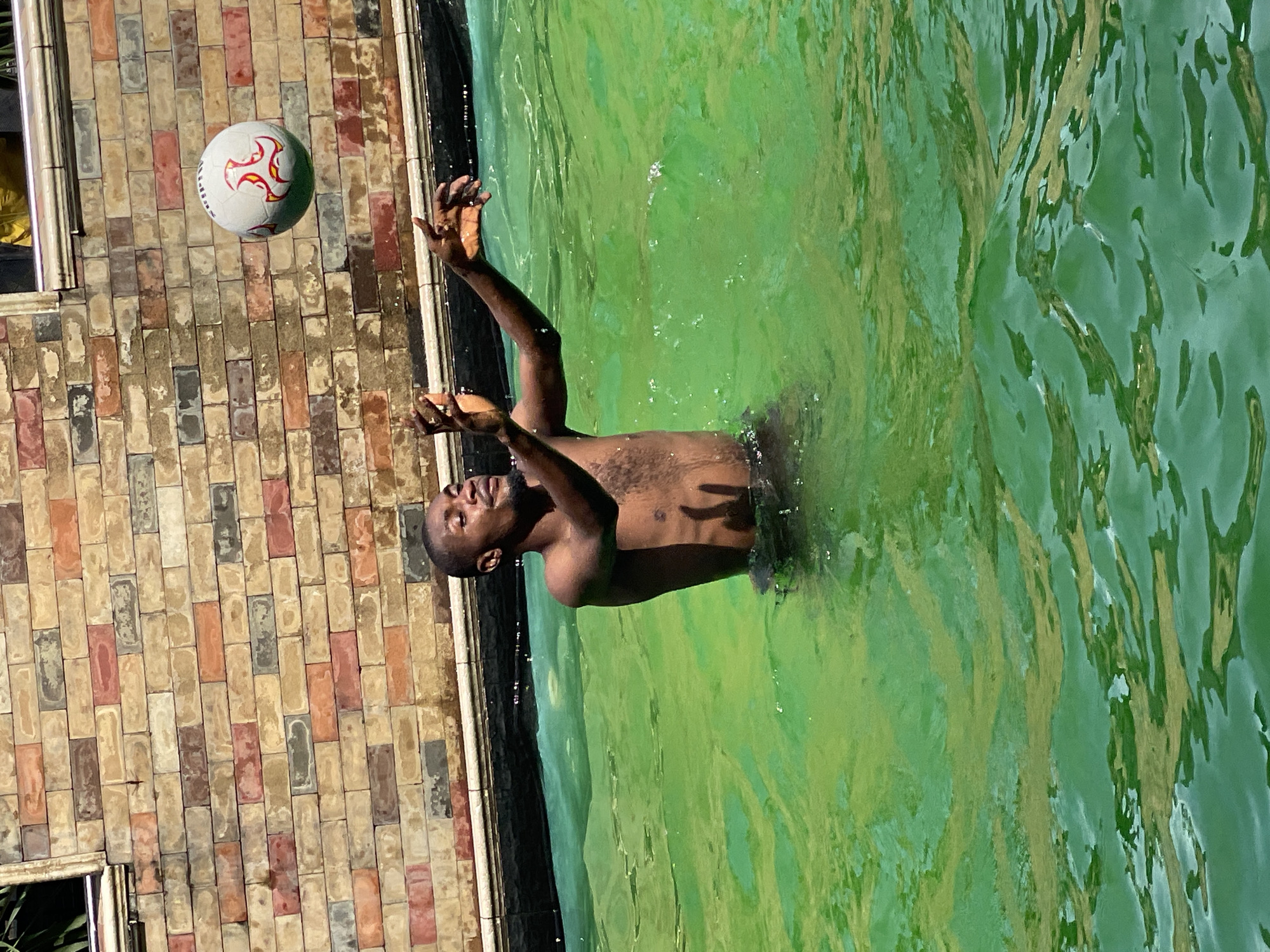 A person is playing with a ball in a swimming pool. The person is a Black male, shirtless, with short hair, and appears to be in his late teens or early twenties. He is standing in green water, with his arms outstretched and hands up, as if catching or throwing a ball. A white soccer ball with red markings is in the air, above and in front of him. The pool is surrounded by a brick wall, made of rectangular bricks in various shades of red, brown, and gray. The lighting suggests it is daytime, with the sun casting shadows and highlighting the ripples in the water. The person's expression is one of concentration and enjoyment. The setting appears to be an outdoor swimming pool, possibly at a residential property or a recreational facility.