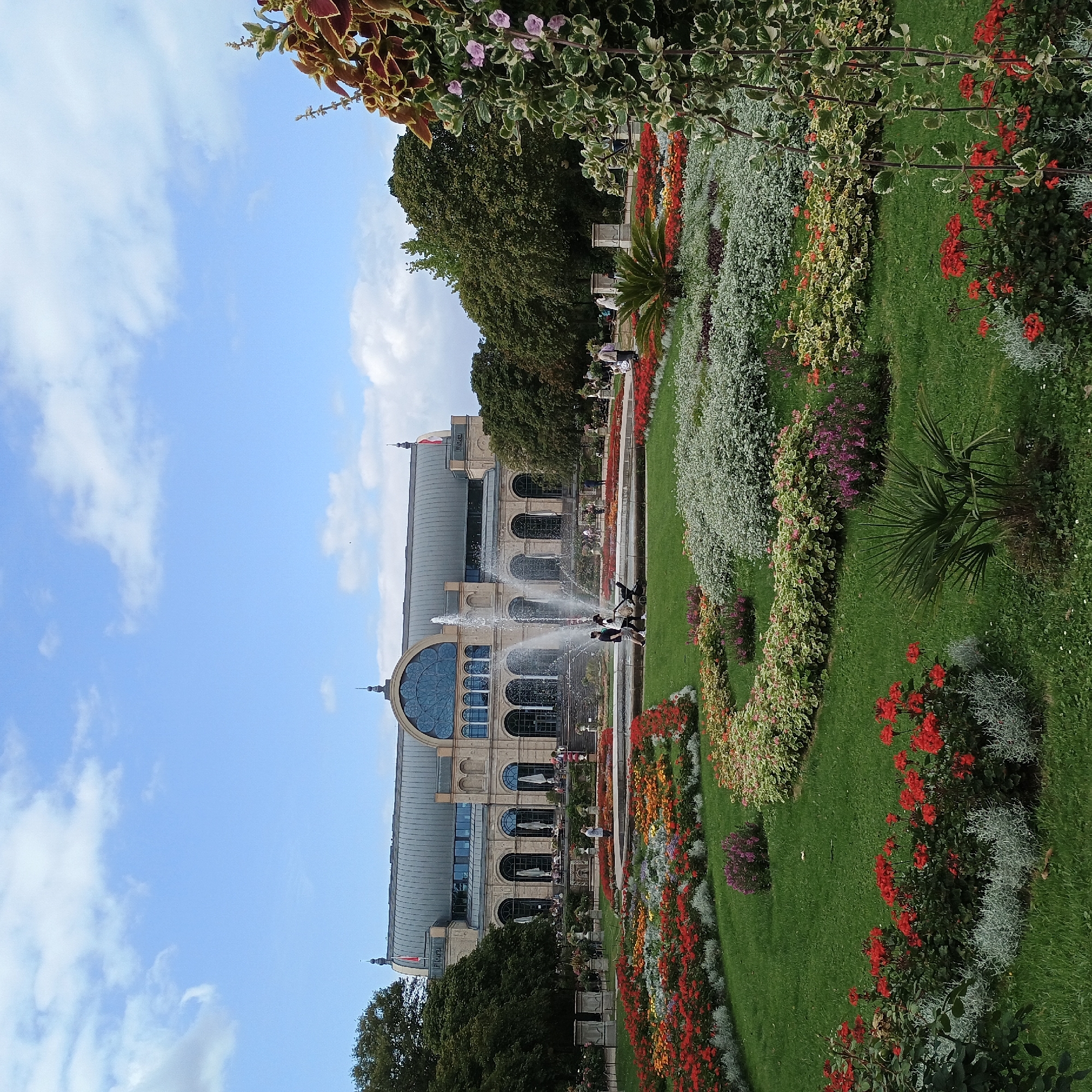 The image depicts a scenic view of the Flora botanical garden in Cologne, Germany. In the background, a grand building with a curved, grey roof and large arched windows stands prominently. The building's facade is light-colored stone, featuring classical architectural elements. In front of the building, a multi-tiered fountain sprays water upwards, creating a dynamic visual element.
The foreground is dominated by a meticulously landscaped garden filled with a vibrant array of flowers. Lush green grass serves as the base for numerous flower beds, showcasing a variety of colors including reds, oranges, yellows, purples, and whites. Silver-grey foliage plants are interspersed among the colorful blooms, adding texture and contrast. A small palm-like plant is situated in the middle of the green lawn.
Several people are visible in the scene. A group of three individuals, including a person with a stroller, walks near the fountain. Other figures are scattered throughout the garden and on the terrace of the building, suggesting a public park setting. Trees with dense green foliage flank the building and the garden, contributing to the overall natural beauty of the location. The sky above is a clear blue, dotted with fluffy white clouds. The overall impression is one of a well-maintained and inviting public space.