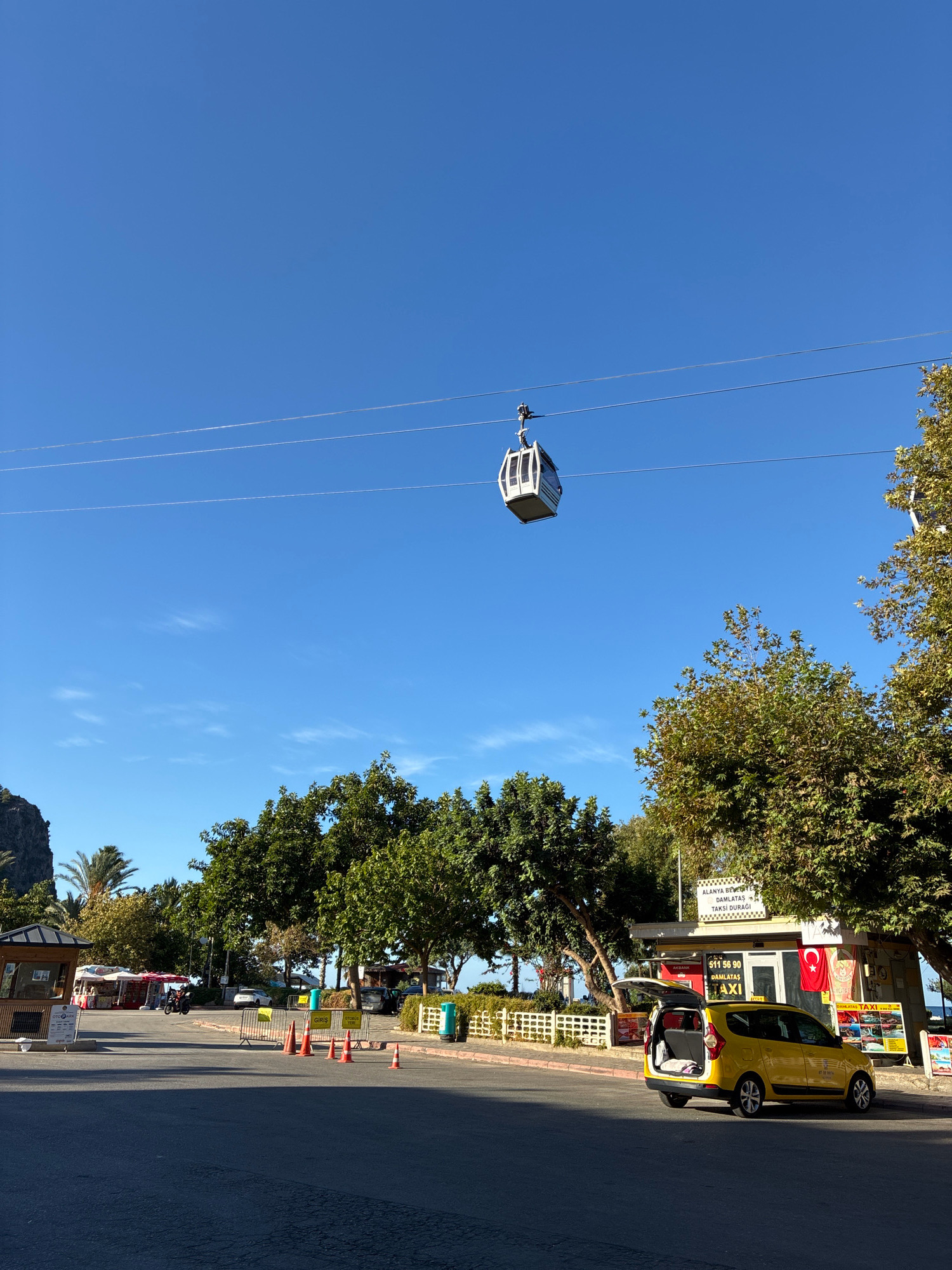 A yellow taxi with its trunk open is parked on the side of a street in Alanya, Turkey. A cable car is suspended in the sky above a tree-lined area. The scene appears to be in a public space, possibly a tourist area, given the presence of kiosks and signage. The sky is clear and blue, suggesting a sunny day. The image also captures elements of the urban environment, including a road, trees, and a building.