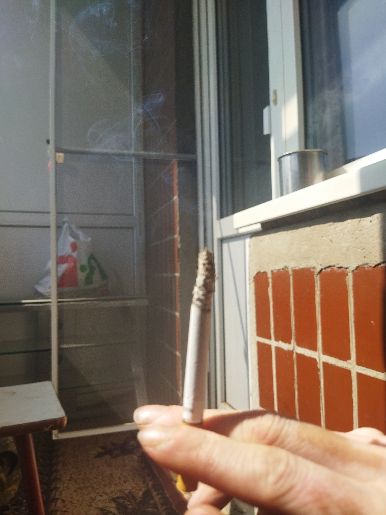 A person's hand holds a lit cigarette, with smoke curling upwards. The scene appears to be on a balcony, indicated by the presence of a window and tiled wall. A table and a bag on a shelf are visible behind the person's hand. The presence of the bag and the style of window suggest that the location is likely a residential area. There is no information in the image to confirm its location. It is not possible to confirm the location as Kramatorsk, Ukraine.