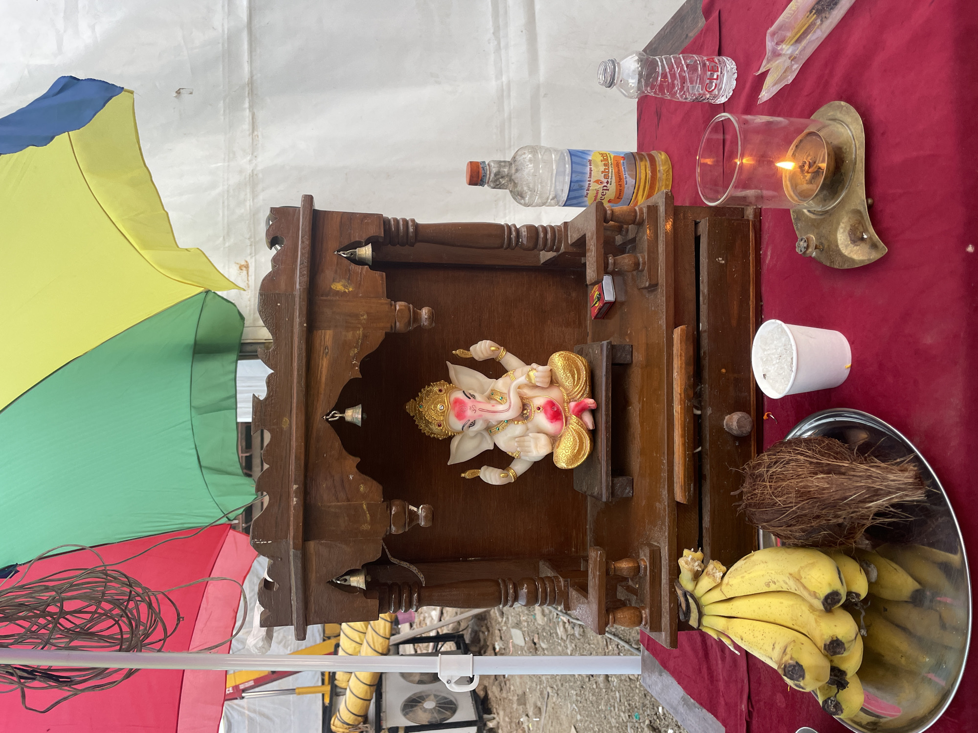 The image shows a Hindu shrine setup outdoors, likely in Mira-Bhayander, India. The shrine is made of dark wood and houses a statue of Ganesha, adorned with gold and pink details. Offerings are arranged around the shrine on a red surface, including bananas, a coconut, a cup of white granular material, and bottles of water and oil, along with a lit candle. To the left of the shrine is a multi-colored umbrella and a coil of wires. An air conditioning unit can be seen in the background. The ground appears to be concrete.