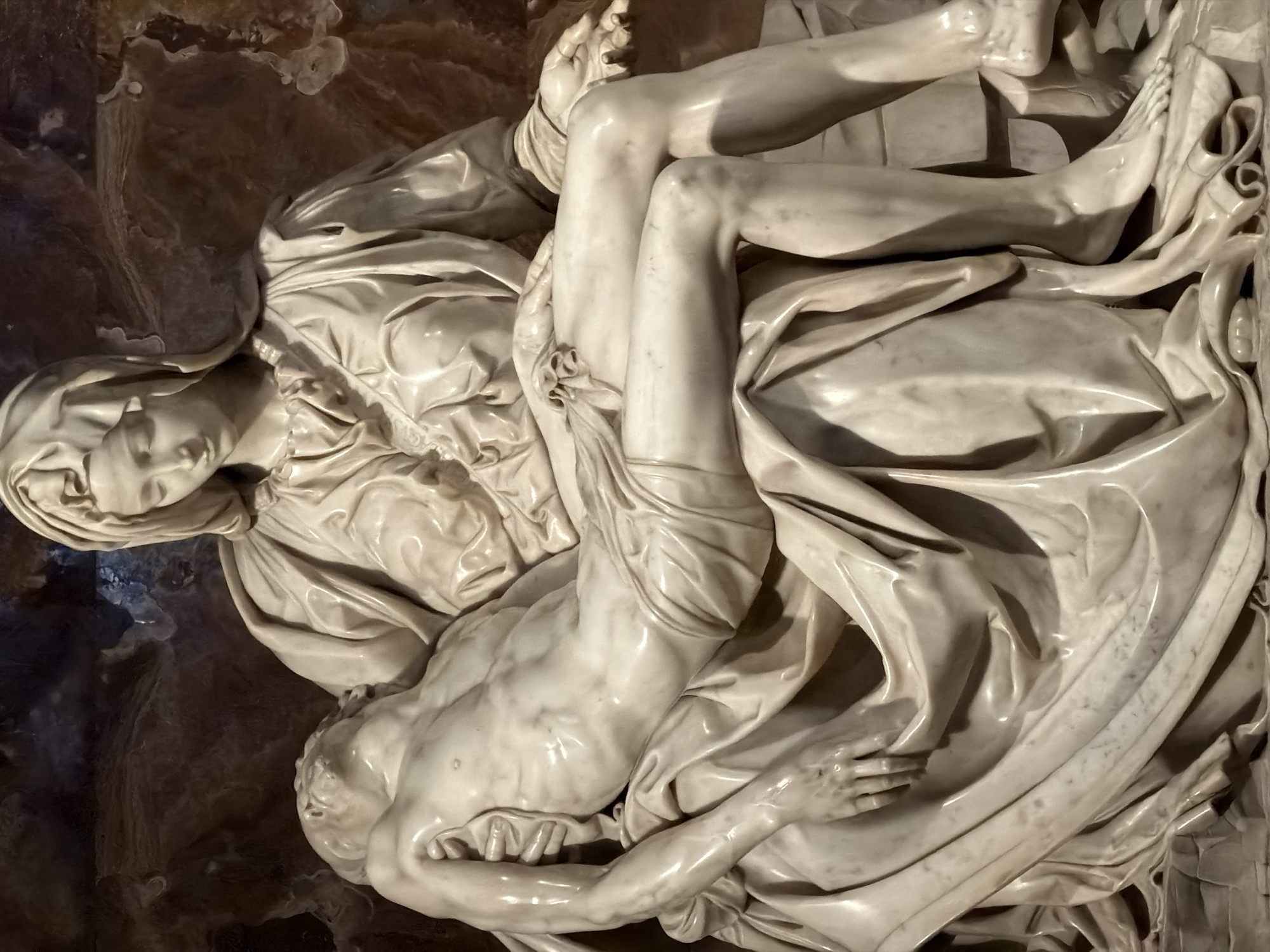 The image depicts the marble sculpture "Pietà" by Michelangelo, located in St. Peter's Basilica in Città del Vaticano, Vatican City. The sculpture features two primary figures carved from white marble with a polished finish, exhibiting subtle veining and reflections from ambient light. The central figure is the Virgin Mary, seated with a somber expression, her head slightly bowed, gazing downwards. She is draped in voluminous, flowing robes and a veil that create deep, realistic folds and shadows. Across her lap lies the supine, lifeless body of Jesus Christ. His head is tilted back, his eyes closed, and his mouth slightly ajar. His torso is visible, revealing anatomical detail and a lean physique, with a small loincloth draped around his waist and upper thighs. His right arm rests across Mary's left thigh, while his left arm is gently positioned over his midsection. His legs are bent, and his feet, showing nail wounds, are visible at the bottom right. Mary's left hand is open, palm up, in a gesture of acceptance or offering, while her right hand supports Christ's body near his armpit. The entire sculpture rests against a mottled, dark reddish-brown background, likely a stone wall or marble cladding within the basilica.