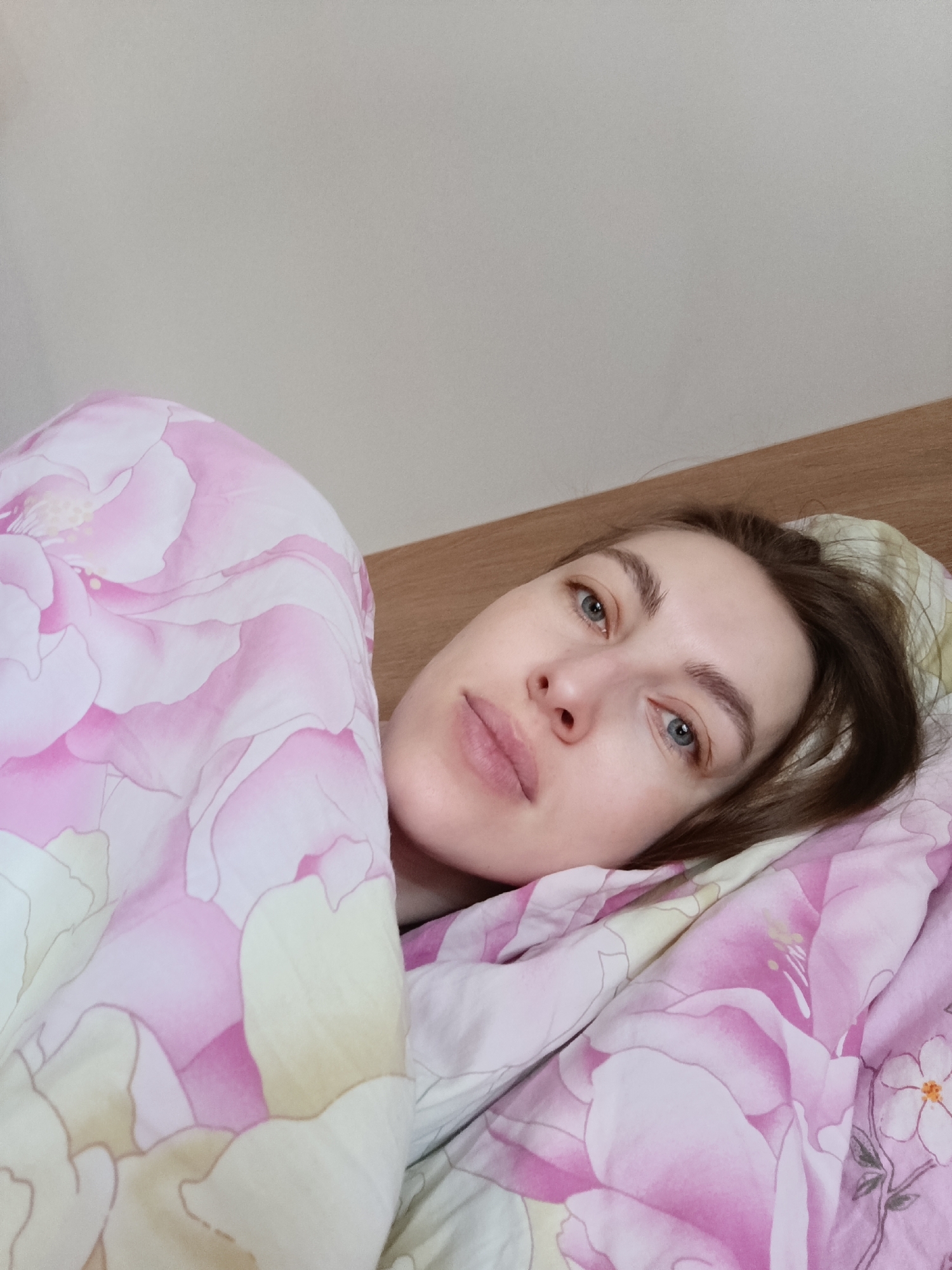 A woman lies in a bed covered with floral patterned sheets, partially covered by a floral patterned blanket. Her head rests on a pillow. She has fair skin, blue eyes, and light brown hair. She is looking directly at the camera. The headboard of the bed is made of wood. The wall behind the bed is pale. The image was reportedly taken in the city of Chaikovsky, Russia.