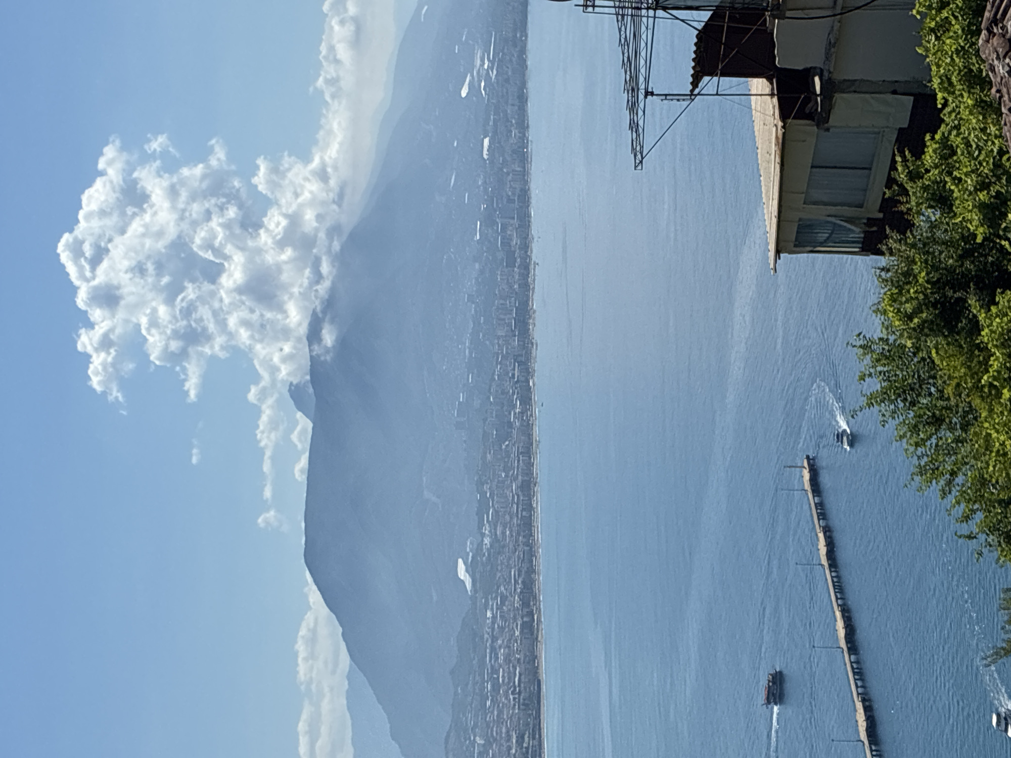 The image depicts a scenic view of Alanya, Turkey, with a focus on the coastline, the Mediterranean Sea, and the Taurus Mountains. In the foreground, a building with a corrugated metal roof and windows is partially visible, situated on a hillside covered with green foliage. A large, wooden pier extends into the bright blue sea from the left side of the frame. Two boats are visible in the water; one is a small, dark-colored vessel moving away from the pier, and another, a white motorboat, is creating a wake as it travels towards the pier. In the middle ground, the city of Alanya stretches along the coast, characterized by numerous buildings and structures. The background is dominated by the majestic Taurus Mountains, which are partially obscured by a light haze and dramatic, white cumulus clouds that appear to be forming a plume above the highest peak. The sky is a clear, vibrant blue, with scattered white clouds. The lighting suggests it is a bright, sunny day.