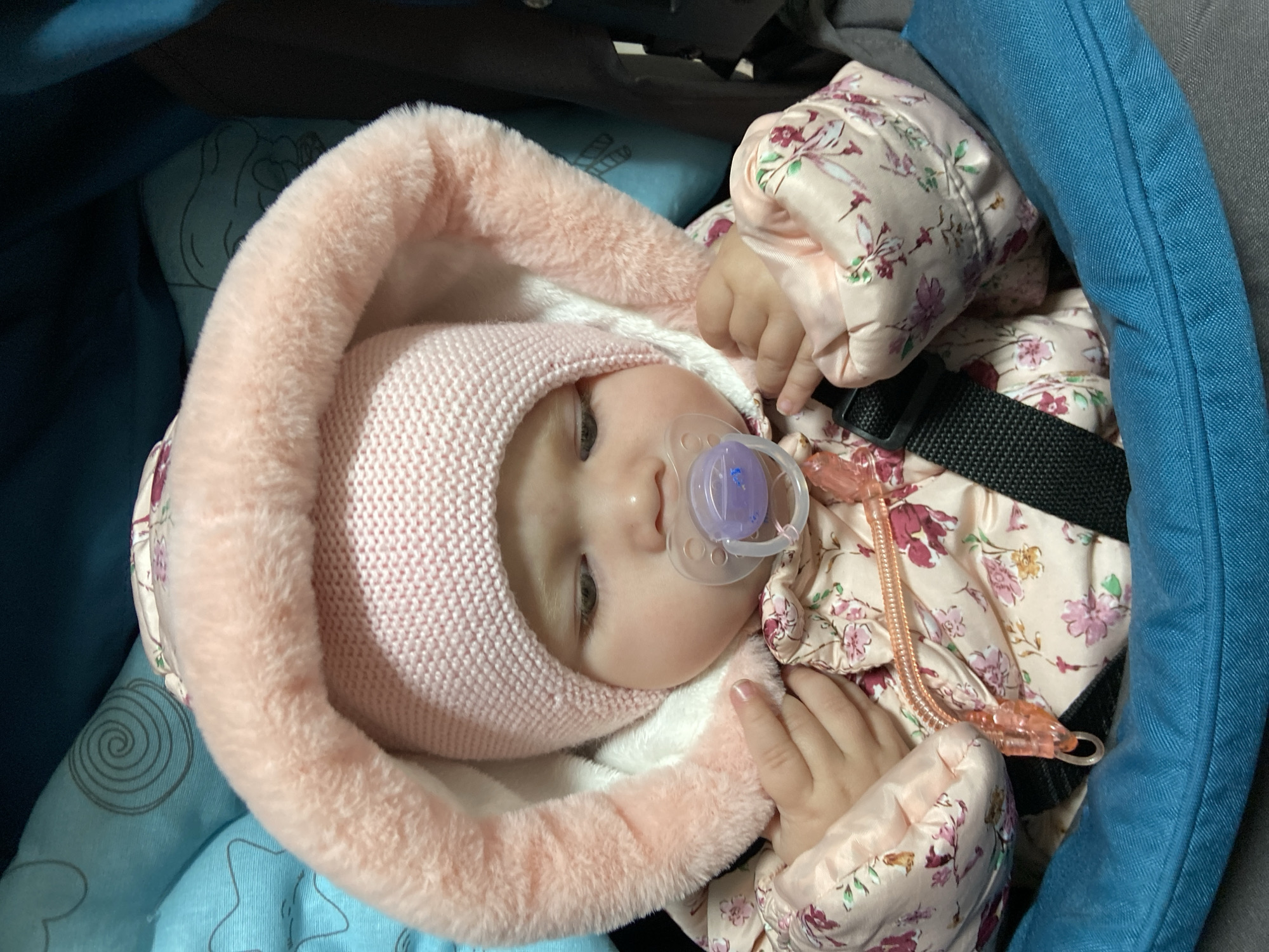 A close-up image depicts an infant seated in a teal-colored stroller or car seat in Фоки, Russia. The infant wears a pink knitted hat and a light pink hooded jacket adorned with a floral pattern of pink and purple blossoms and green leaves. The hood features a white fluffy lining and a pink fur trim. A clear pacifier with a purple nipple is inserted into the infant's mouth. The infant's eyes are cast downwards and to the left. Small hands are visible, one resting near a pink plastic zipper pull on the jacket. The teal fabric of the stroller/car seat contains subtle grey patterns, including swirls and stars, on the inner lining behind the infant's head. A black safety strap is visible across the lower right side of the infant's body.