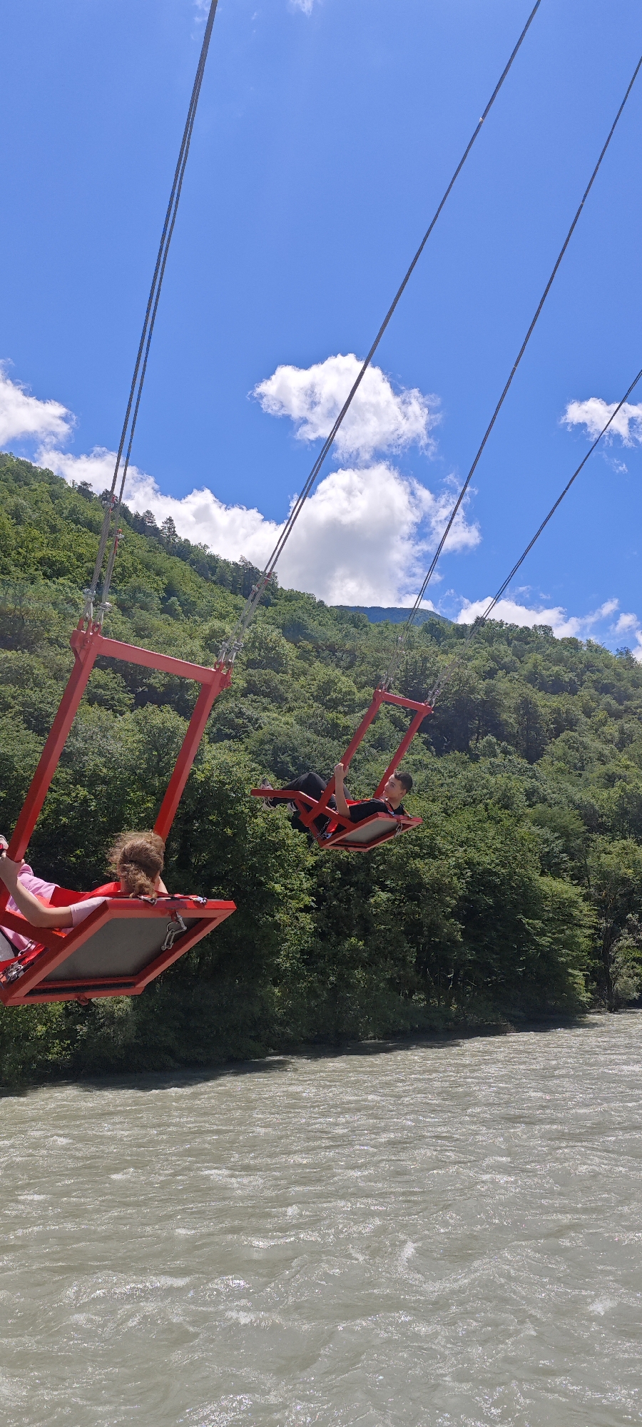 Two individuals are captured mid-air on large, red swings, suspended by thick metal cables high above a turbulent river. The scene is set against a backdrop of steep, densely forested mountains under a bright blue sky dotted with scattered white cumulus clouds.
In the foreground, a person with curly light brown hair, wearing a pink top, is seated in one of the red swings, facing away from the camera and towards the landscape ahead. Their swing appears to be moving, lifted slightly higher than the other. Further back and to the right, another person, appearing male, with dark hair, wearing a black shirt and dark pants, is comfortably reclined in a similar red swing. He is looking towards the left, seemingly enjoying the panoramic view and the thrill of the ride. Both swings are designed with sturdy red frames and flat platforms for seating, secured by harnesses.
Below the swings, a wide river flows with light-colored, foamy water, indicating a swift current. The opposite bank of the river is lined with more dense green trees that extend up the slopes of the mountains. The sunlight is bright, suggesting a clear day. The atmosphere conveys a sense of adventure and exhilaration. There is no visible text in the image. The location is Bzypta, Abkhazia, known for its mountainous terrain and rivers.