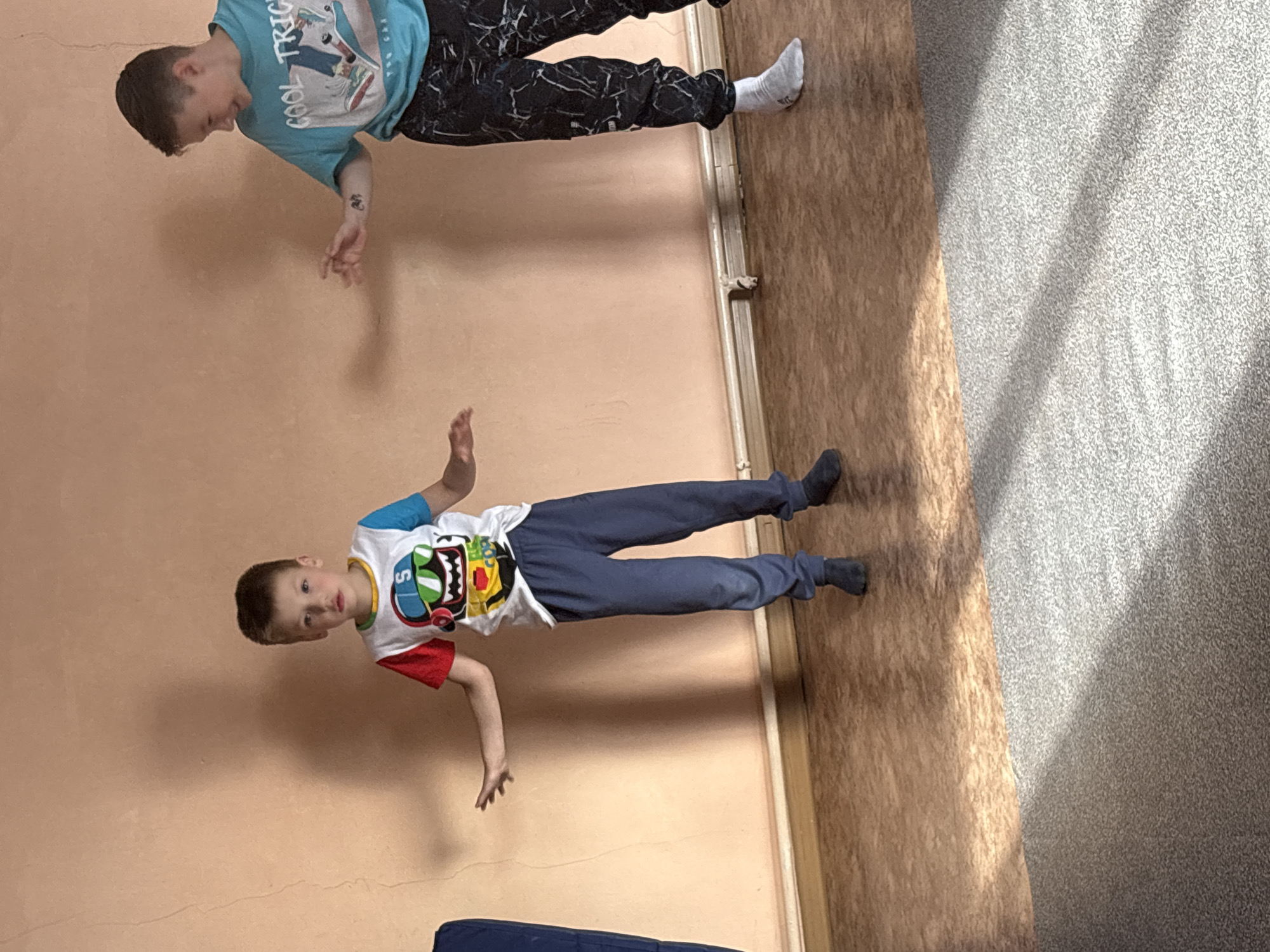The image shows two young boys indoors, appearing to be engaged in some activity. The boy on the left, wearing a colorful t-shirt with a graphic print and blue pants, stands with his arms outstretched, looking towards the camera with a neutral expression. The boy on the right, in a turquoise t-shirt with the text "COOL TRIC" visible and patterned black pants, is turned to his left, looking down and to the side with a slight smile, his left arm extended. A faint tattoo is visible on his left forearm. They are positioned in a room with a peachy-colored wall and a wooden floor transitioning to a grey carpeted area. Sunlight casts shadows on the floor from the left. The location is specified as a room in the city of Chaikovsky, Russia.