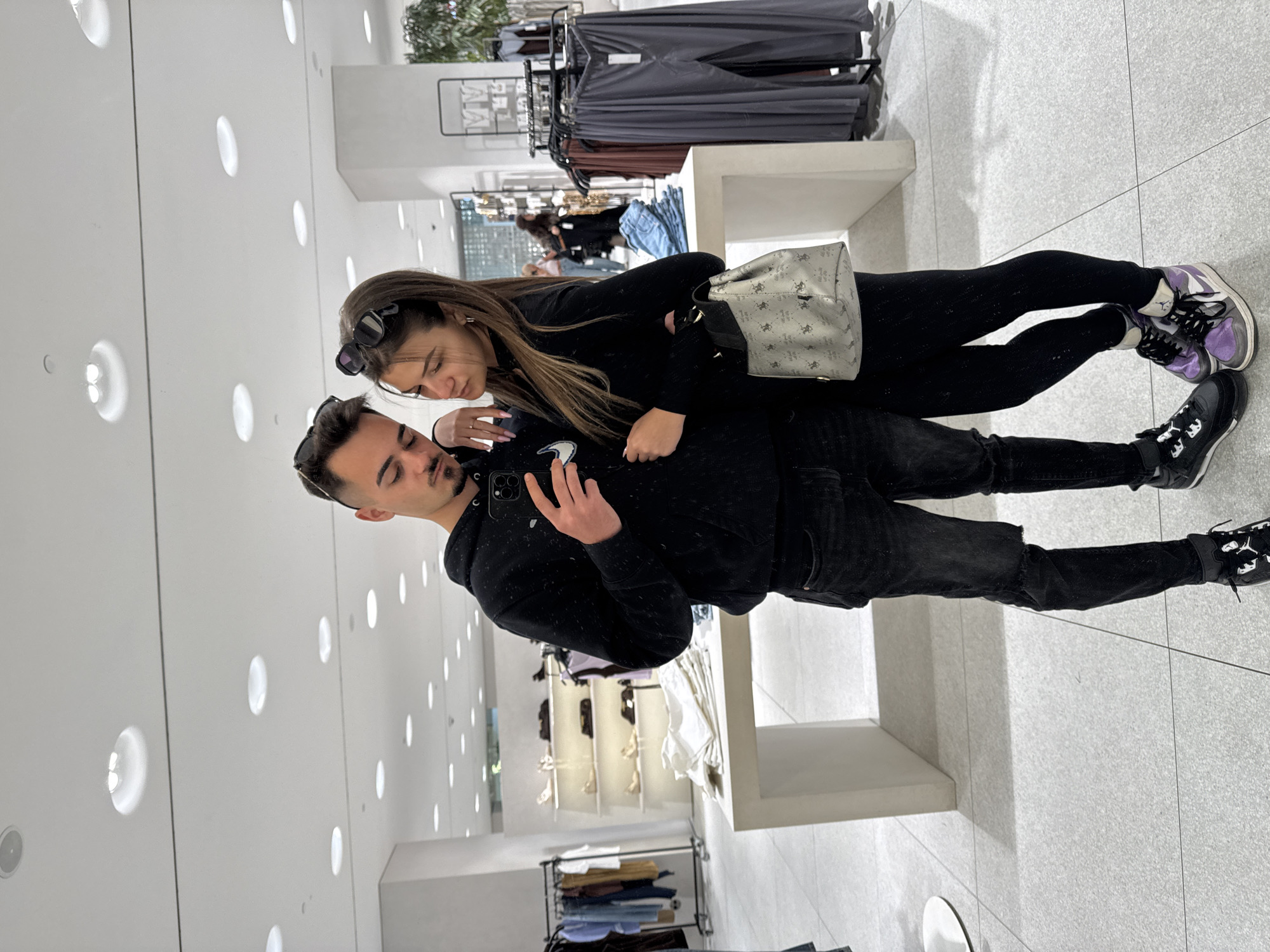 In a brightly lit retail store with recessed circular ceiling lights and neutral tile flooring, a man and a woman are posing for a selfie. The man, dressed in a black hoodie and black jeans, is holding a smartphone in his right hand to take the photo. He has dark hair and is wearing sunglasses perched atop his head. The woman, standing slightly behind and to the side of the man, is wearing black leggings and a black top. She has long brown hair and sunglasses also on her head. She is carrying a light-colored patterned shoulder bag. Clothes racks with garments can be seen in the background, suggesting a clothing store setting. The location is possibly in Sofia, Bulgaria, but there are no specific elements that confirm this.