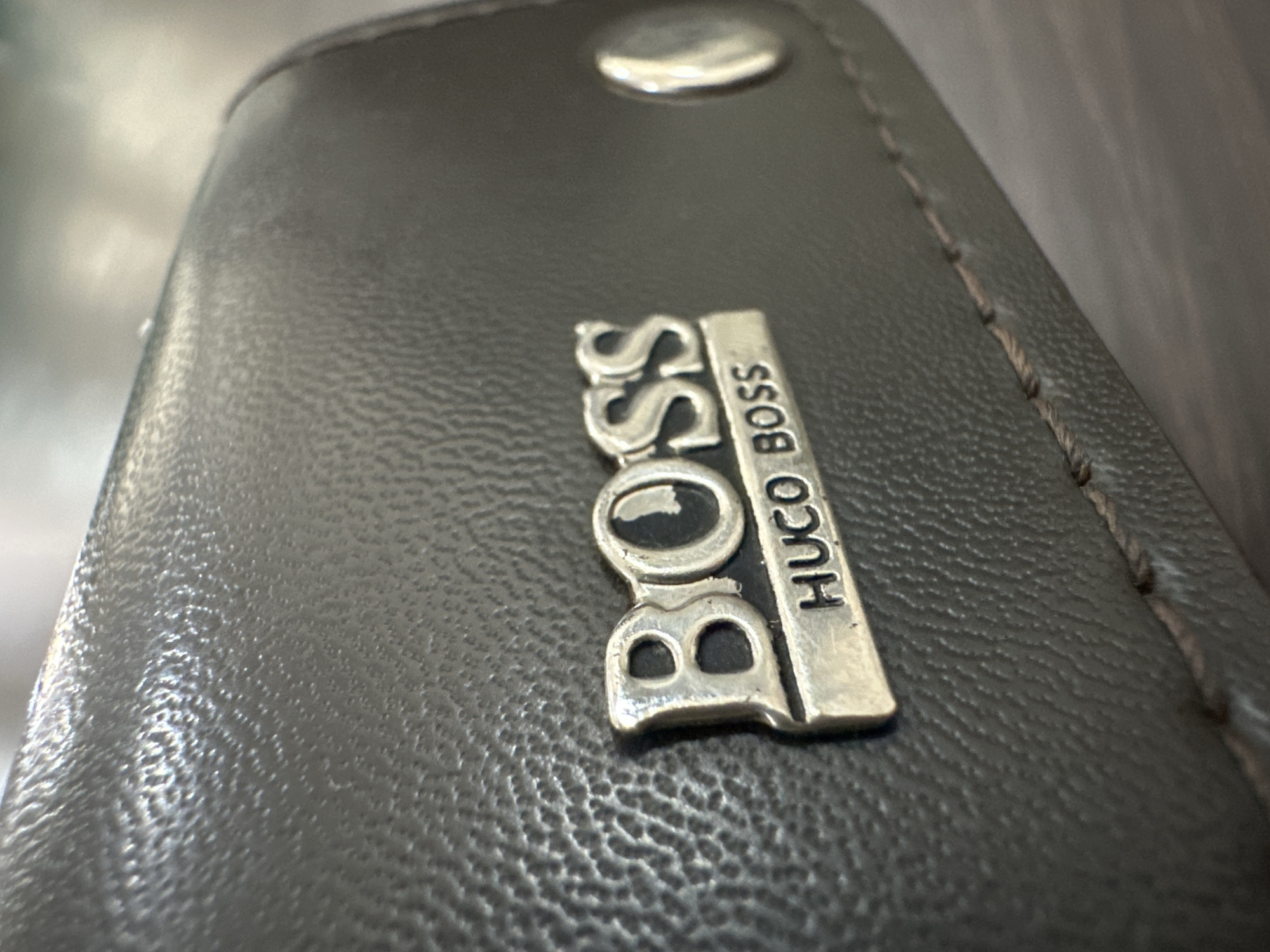 The image is a close-up, eye-level shot of a dark grey, textured leather object, likely a wallet or small bag. A silver-colored metal logo is prominently displayed on the surface. The logo consists of two parts: the word "BOSS" in large, raised, metallic letters, and below it, a smaller rectangular plate with the words "HUGO BOSS" inscribed. The leather has a subtle, pebbled texture, and stitching is visible along the bottom edge of the object, suggesting it is a manufactured item. There is a silver stud or button visible on the right side of the object, partially out of focus. The background is blurred and appears to be a neutral, indoor surface, possibly a wooden table, suggesting the object is placed on a flat surface. The lighting is soft, highlighting the metallic sheen of the logo and the texture of the leather. There are no people or animals in the image. The image does not contain any discernible information about the location in the city of Bulakrejo, Indonesia.